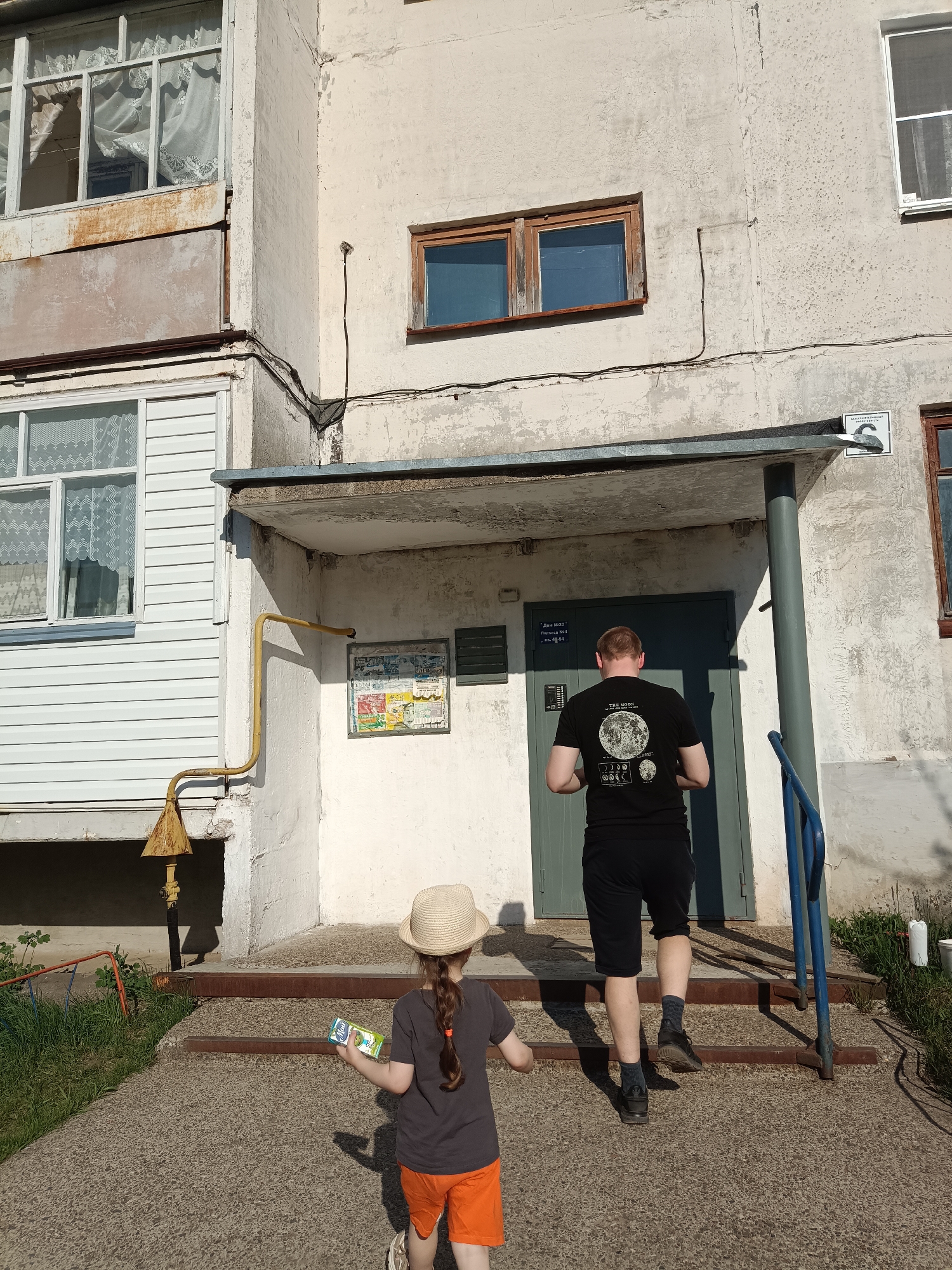 A man and a girl approach the entrance of a nondescript building. The building is a weathered, two-story structure with visible signs of aging, including cracked walls and peeling paint. The entrance has a small, overhanging roof supported by pillars. A blue metal handrail is on one side of the stairs. The man, seen from the back, wears a black t-shirt with a moon graphic, black shorts, dark socks, and sneakers. The girl, slightly ahead of him, wears a straw hat, a gray t-shirt, and orange shorts. She holds a small juice box. The environment includes a patch of grass and some vegetation on either side of the entrance path. A yellow gas pipe runs along the side of the building. A sign is affixed to the wall near the entrance. The building's exterior and architectural style suggest it is in Noviy, Russia. The interaction is between the man and the girl, implying they are entering the building together.