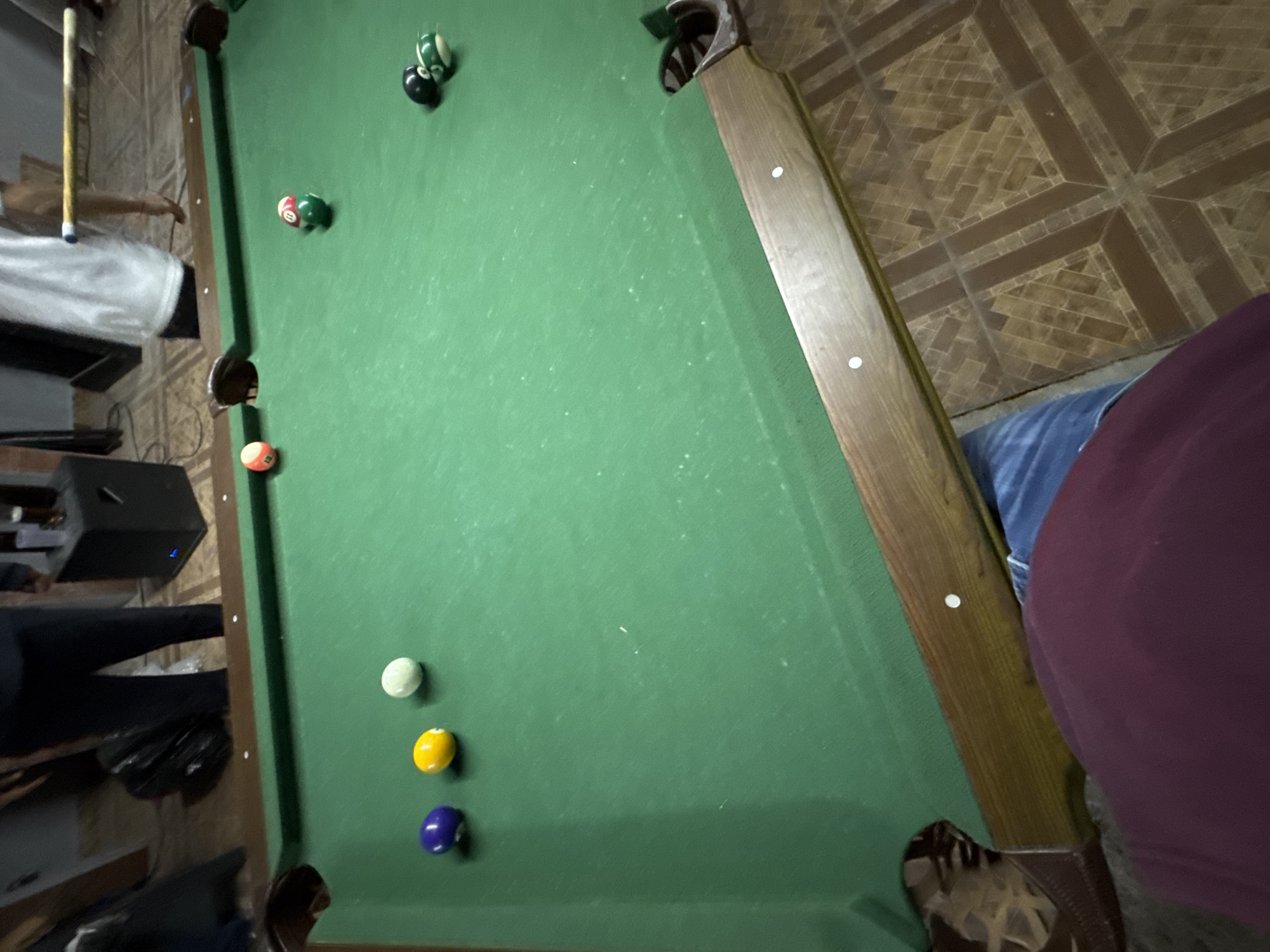 A medium-angle, slightly tilted photograph depicts a pool table in what appears to be an indoor setting, possibly in Heroica Puebla de Zaragoza, Mexico. The table is covered with green felt and has several billiard balls scattered across its surface. A cluster of balls is located in the upper section of the table near a corner pocket. Lower down, near the bottom edge, are three additional balls lined up. A person in a dark colored shirt and blue jeans is positioned on the right side of the frame, partially obscuring the table's edge. Another person standing on the left side of the frame with a cue stick. The surrounding environment includes tiled flooring and what may be furniture or other equipment. Overall, the scene seems to capture a moment during a pool game.