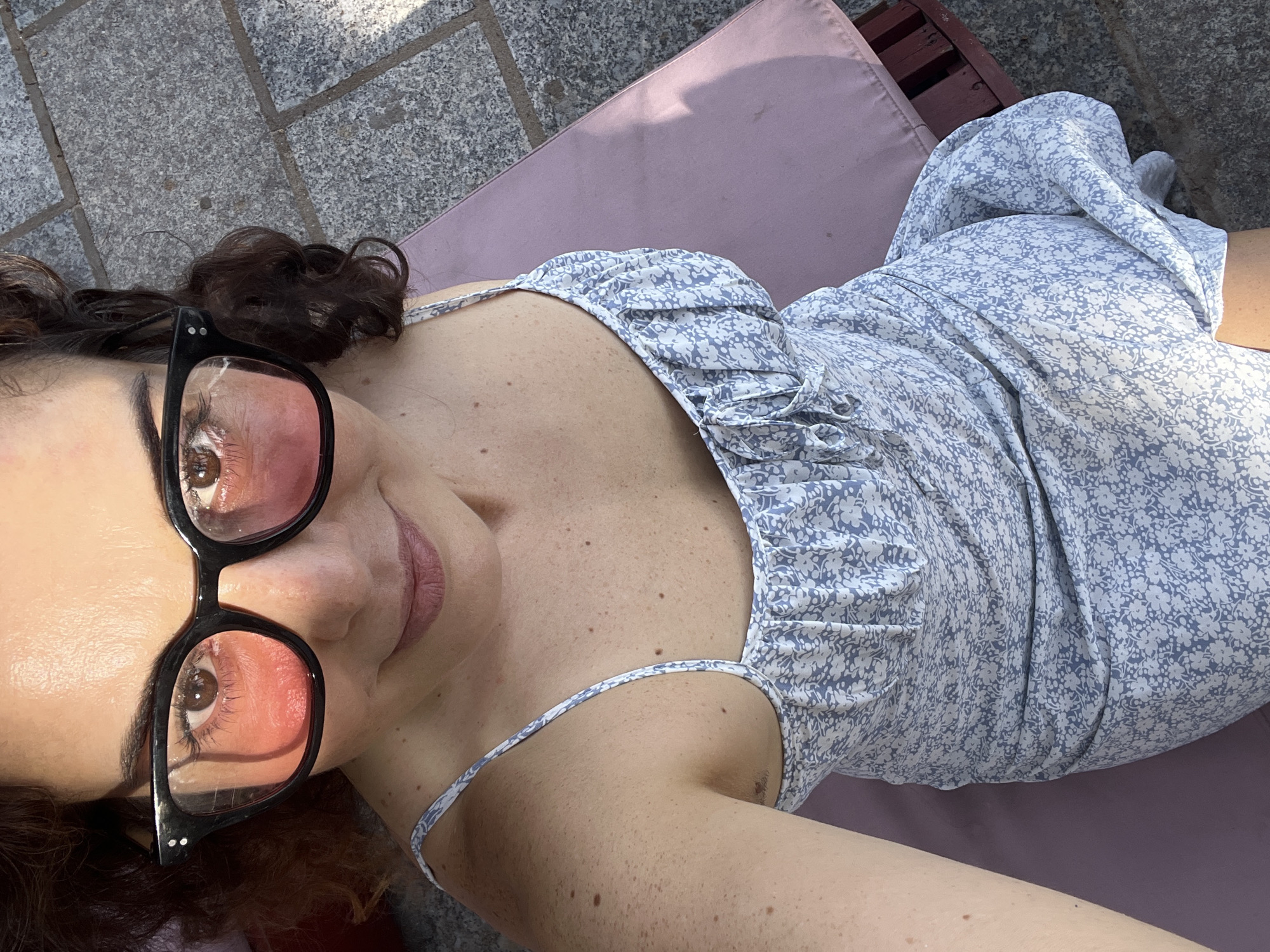 A woman with curly brown hair, wearing black-rimmed glasses with pink lenses and a blue and white floral print camisole dress, lies on a pink lounge chair outdoors. The background shows a paved area with grey flagstones. The scene appears to be taken during daylight, with sunlight casting shadows. The woman is looking up at the camera, with her head tilted. Her skin has visible freckles and she has light pink lipstick on. The dress has a ruffled neckline with drawstrings. She is positioned on the lounge chair in a reclined manner, with her right arm visible.