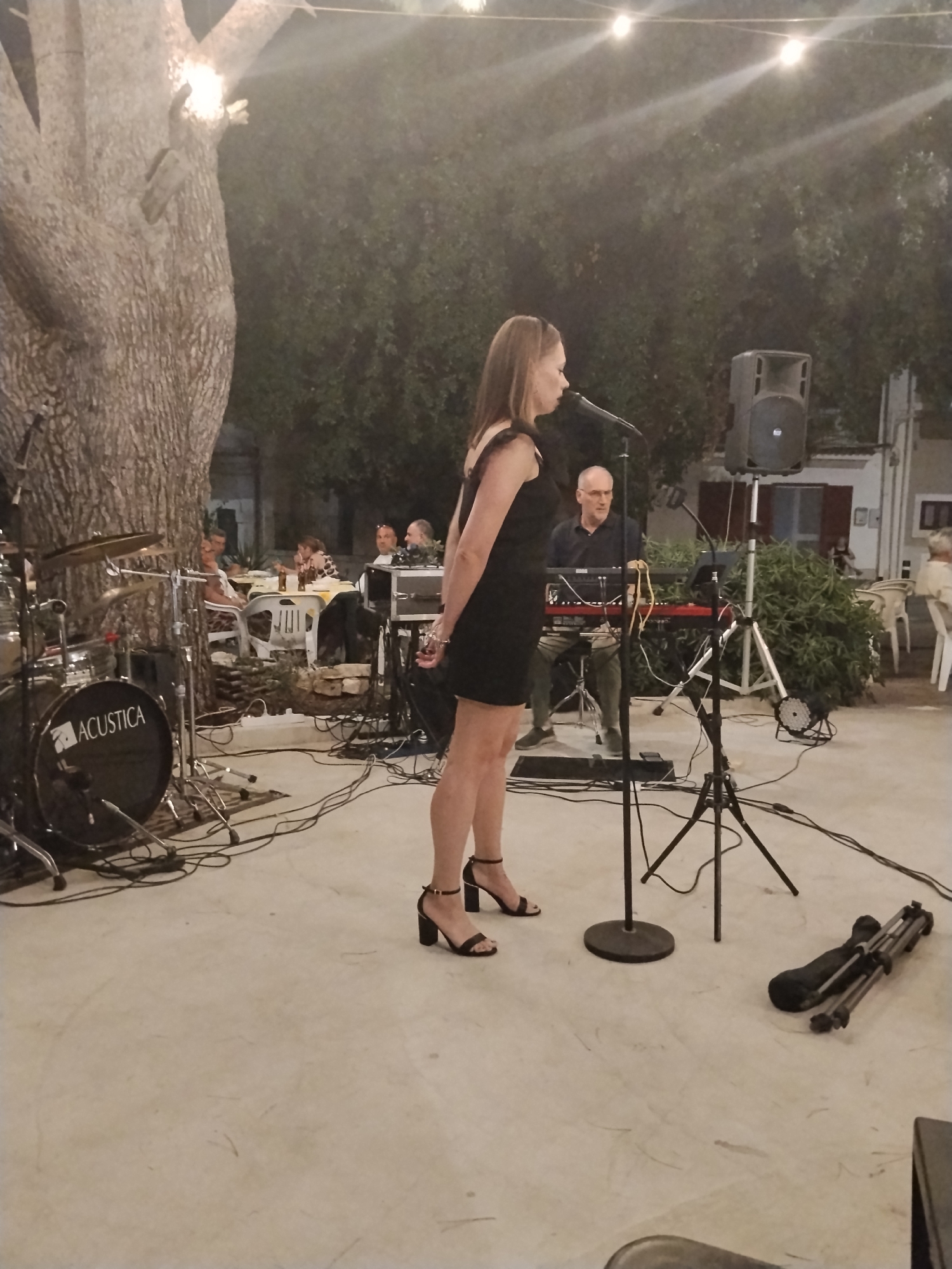 The image depicts an outdoor evening performance. In the foreground, a woman in a black dress and heeled sandals stands in profile, facing a microphone. To her left, a drum set with the "ACUSTICA" logo is visible. Further back, a man wearing glasses and a dark shirt plays a red keyboard. Other individuals are seated at tables in the background, some with bottles of beer. The scene is illuminated by string lights and the ambient light suggests it is dusk or early evening. The location is identified as Toritto, Italy.