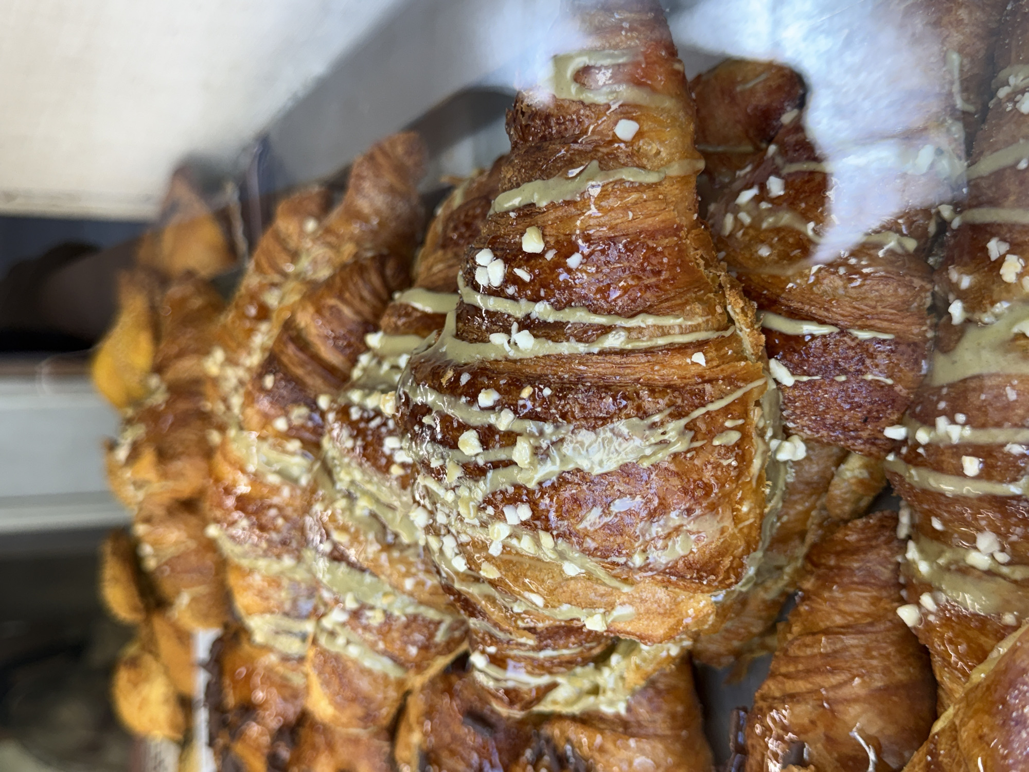 A close-up, slightly elevated shot captures a dense display of freshly baked croissants, likely in a bakery or cafe in Unknown, Unknown. The croissants are predominantly golden to deep amber brown, exhibiting a glossy, lacquered finish from a glaze. Many are generously drizzled with a light green, viscous icing or sauce, possibly pistachio-flavored, and uniformly sprinkled with small, irregularly shaped fragments of light-colored nuts, likely chopped pistachios or almonds. The croissants are stacked several layers deep, filling the majority of the frame, with the foreground showing sharp detail and the background progressively blurring into soft-focused shapes of more pastries and indistinct elements of the interior. A noticeable glare or reflection from a transparent surface, likely a display case, is visible in the lower right portion of the image. The ambient lighting accentuates the shine on the pastries.