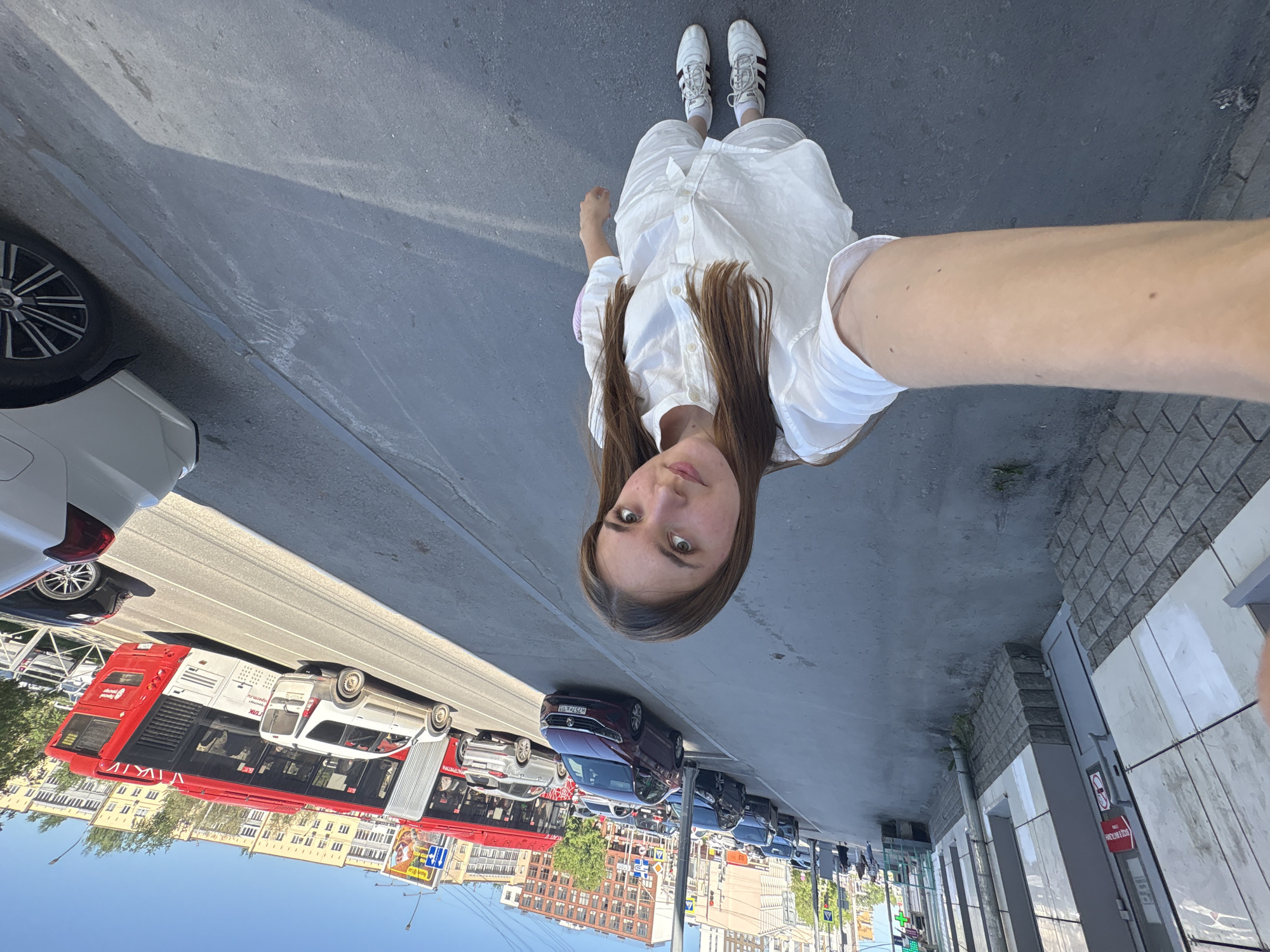 The image shows a woman taking a selfie on a sidewalk in Perm, Russia. She is wearing a white button-down shirt, white pants, and white sneakers with black stripes. The background features a street with cars, buses, buildings, and street signs characteristic of the urban environment. The road shows several vehicles, including a distinctive red and white double-decker bus. The buildings appear to be of an older architectural style, common in Perm's central districts. The sky is clear and blue.