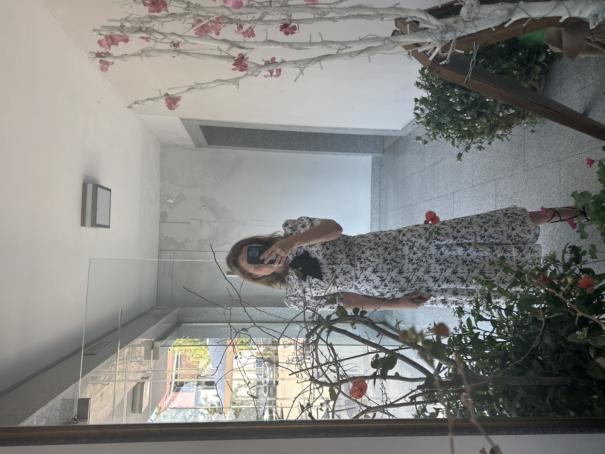 A woman takes a mirror selfie in a hallway. She is wearing a white dress with a black floral pattern, and a black bow at the neck. The hallway has white walls, grey tiled floors, and a large mirror. There are plants and decorative branches in the foreground and to the right. Outside a large window, there are trees and buildings visible.