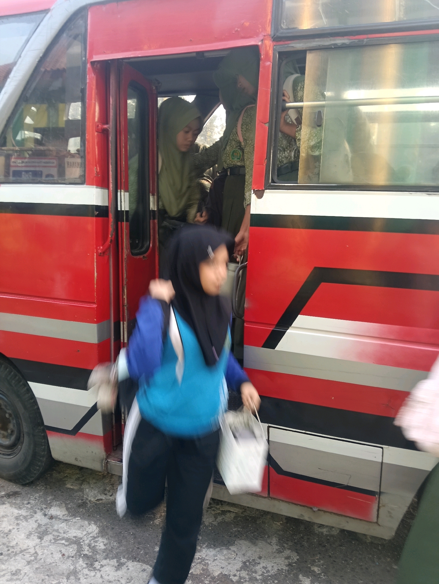 A blurry, eye-level medium shot captures a red and white bus with black and gray stripes in Adinuso, Indonesia. A young girl in a black hijab and blue uniform is running out of the bus with a white bag in her right hand, her left hand holding a light-colored object. She is moving away from the bus and towards the right side of the frame. In the background, another girl in a green hijab and uniform is visible inside the bus, and a third person, also in a green uniform, stands near the doorway. The bus has its doors open, revealing the interior and passengers. The ground appears to be a rough, gray surface. The lighting suggests it is daytime.