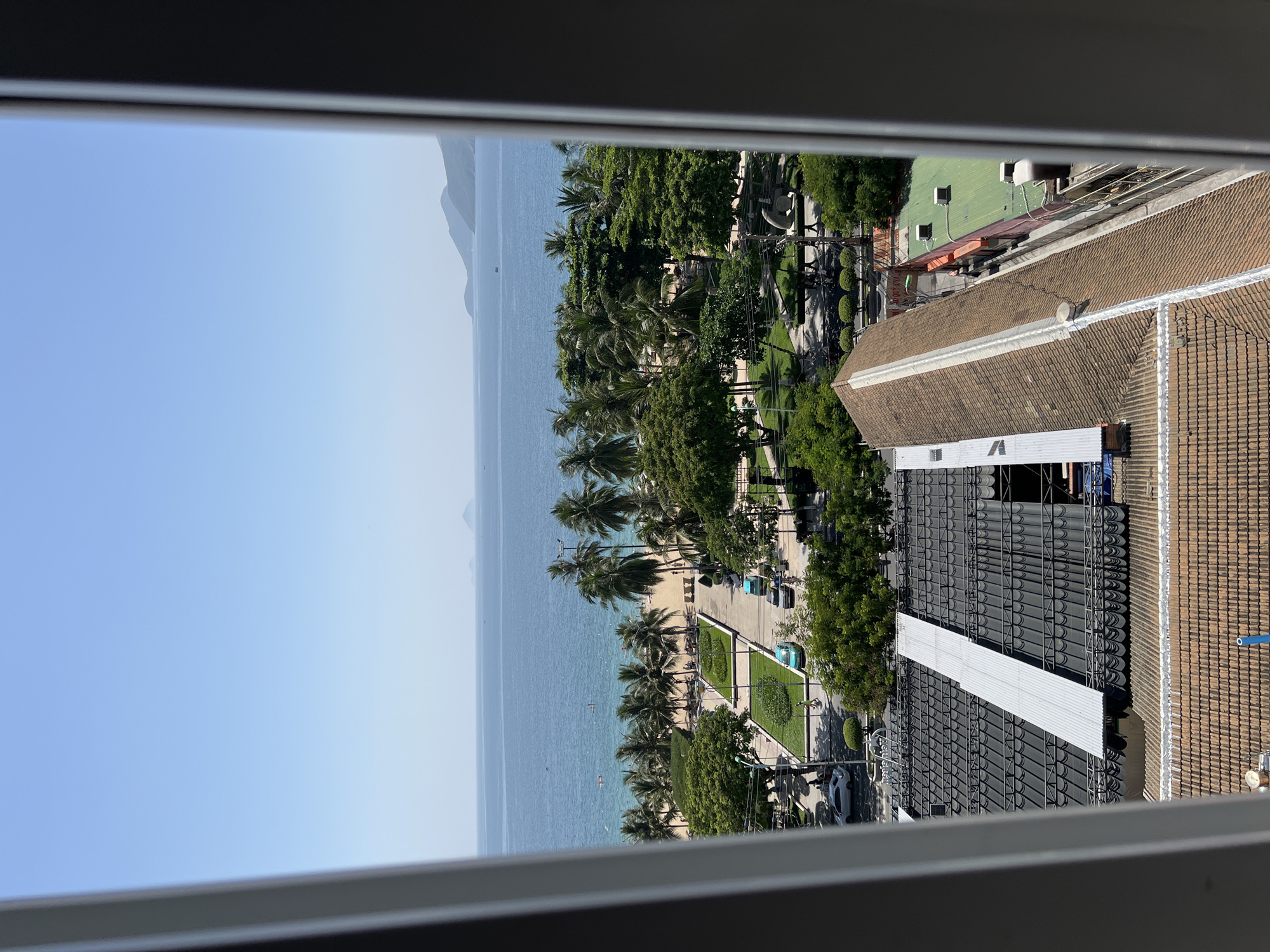 The image presents a view of Nha Trang, Vietnam, from a high vantage point, framed by the edges of a window. In the foreground, the roofs of buildings are visible, with one roof covered in brown tiles and another, larger one, featuring dark gray corrugated metal roofing with a long, reflective silver panel running diagonally across it, supported by a metal framework. Beyond these buildings, a verdant landscape unfolds, characterized by numerous palm trees and other lush green foliage. This green zone appears to be a park or a landscaped area with manicured lawns and pathways.
Further in the distance, a sandy beach stretches along the coastline, bordered by the clear blue waters of the ocean. Several people are visible on the beach and in the water, suggesting recreational activity. A few boats are scattered on the sea, and in the far background, the undulating shapes of mountains and islands can be seen against the horizon under a bright, cloudless blue sky. The presence of cars on the road and people walking on the pathways in the landscaped area indicate a populated urban environment adjacent to the beach. The overall scene depicts a tropical coastal city on a sunny day.