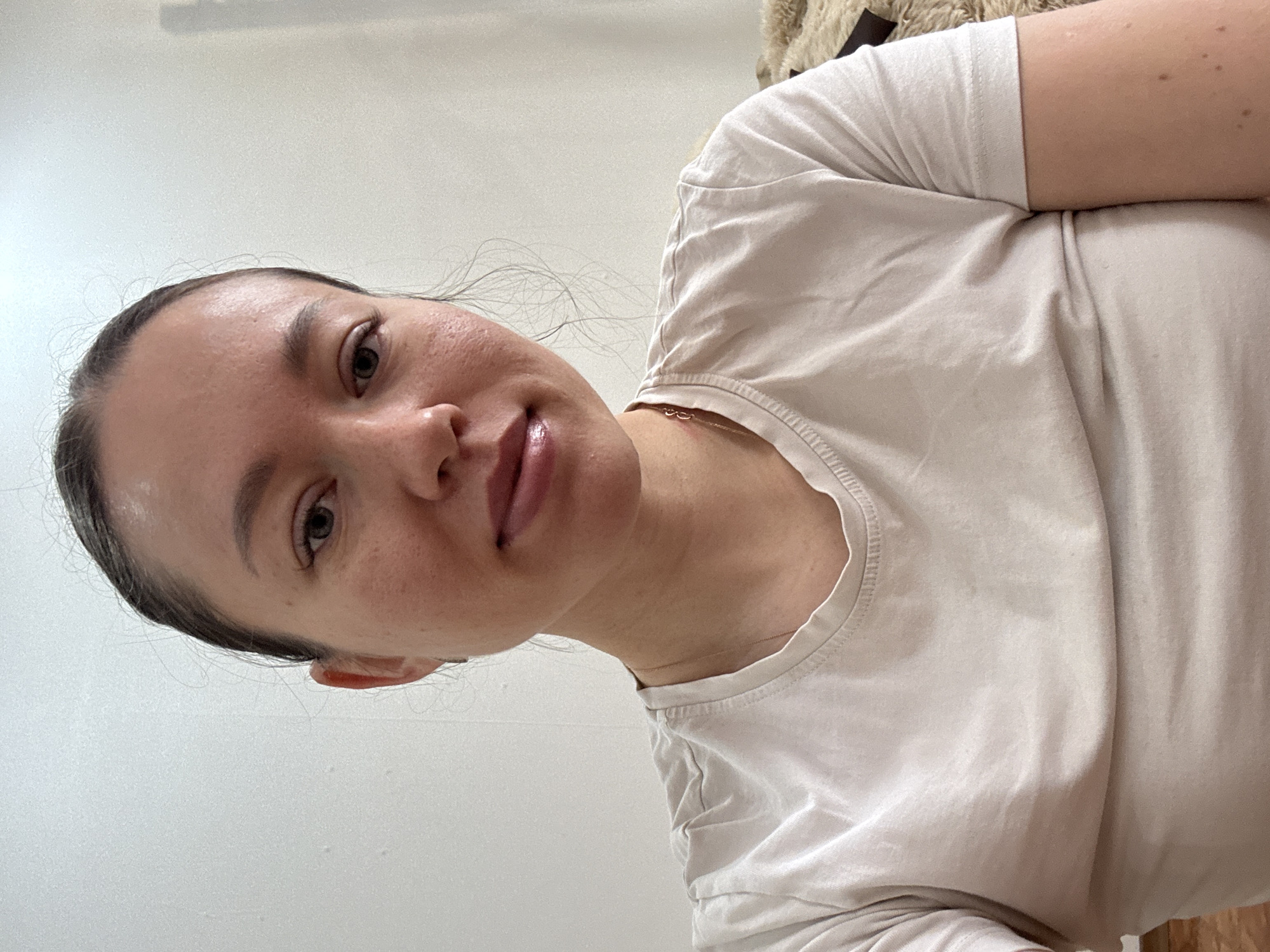 A woman with fair skin, dark hair pulled back, and a neutral expression faces the camera. She is wearing a light-colored T-shirt and a delicate necklace. The background is a plain white wall. No information in the photo indicates that this image was taken in the city of Ферма, Russia.
