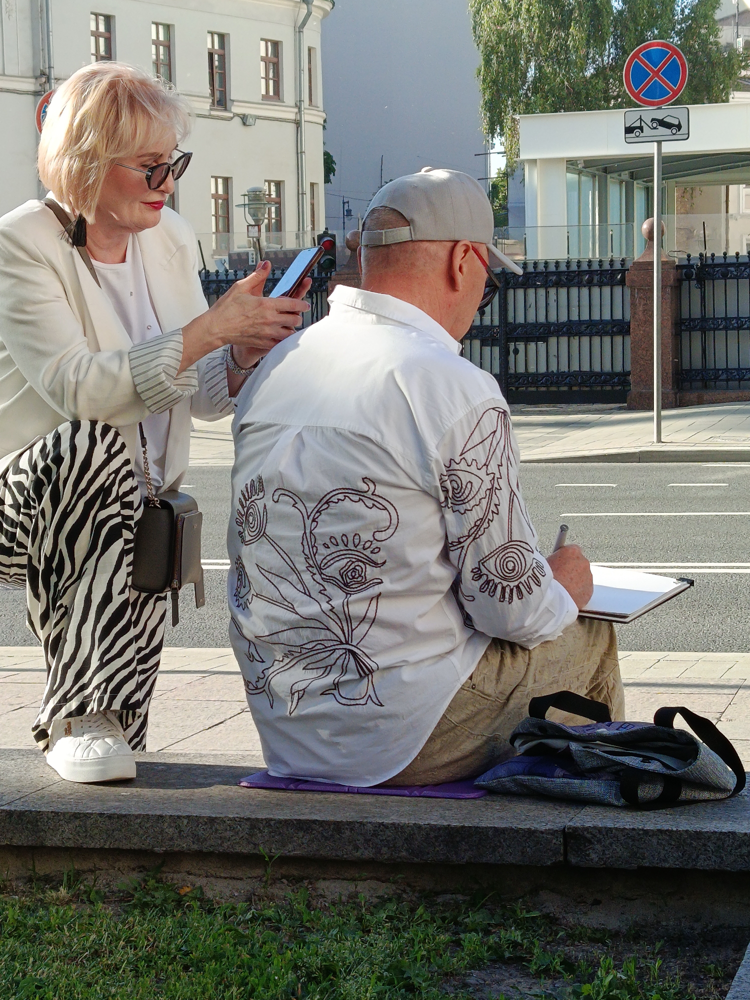 The image depicts an outdoor urban scene on a sunny day, featuring two individuals on a concrete ledge next to a paved area.
On the left, a woman with blonde, shoulder-length hair and sunglasses stands. She wears a white blazer over a white top, black and white zebra-print wide-leg trousers, and white sneakers. A small black shoulder bag with a chain strap hangs at her side. She holds a dark-colored smartphone horizontally with both hands, looking at its screen, appearing to record or photograph.
To her right, a man is seated on the concrete ledge, facing away from the camera. He wears a light grey baseball cap and a white long-sleeved shirt with intricate dark brown embroidery featuring abstract, eye-like patterns on the back and right sleeve. He wears khaki-colored trousers. A purple mat is placed beneath him on the ledge. He holds a pen or marker in his right hand and draws on a white pad or clipboard resting on his lap. Next to him on the ledge is a grey patterned tote bag.
In the background, a paved road with white lane markings is visible. Above the road, a red circular "no stopping" sign with a blue 'X' is mounted on a pole, beneath which is a rectangular grey sign depicting a tow truck. A dark metal fence runs along the street, separating it from buildings. On the left, a light-colored multi-story building with several windows is visible. To the right, behind the fence, another building with glass features is partially seen. Green trees are visible further in the background. The foreground shows a patch of green grass with weeds below the concrete ledge.