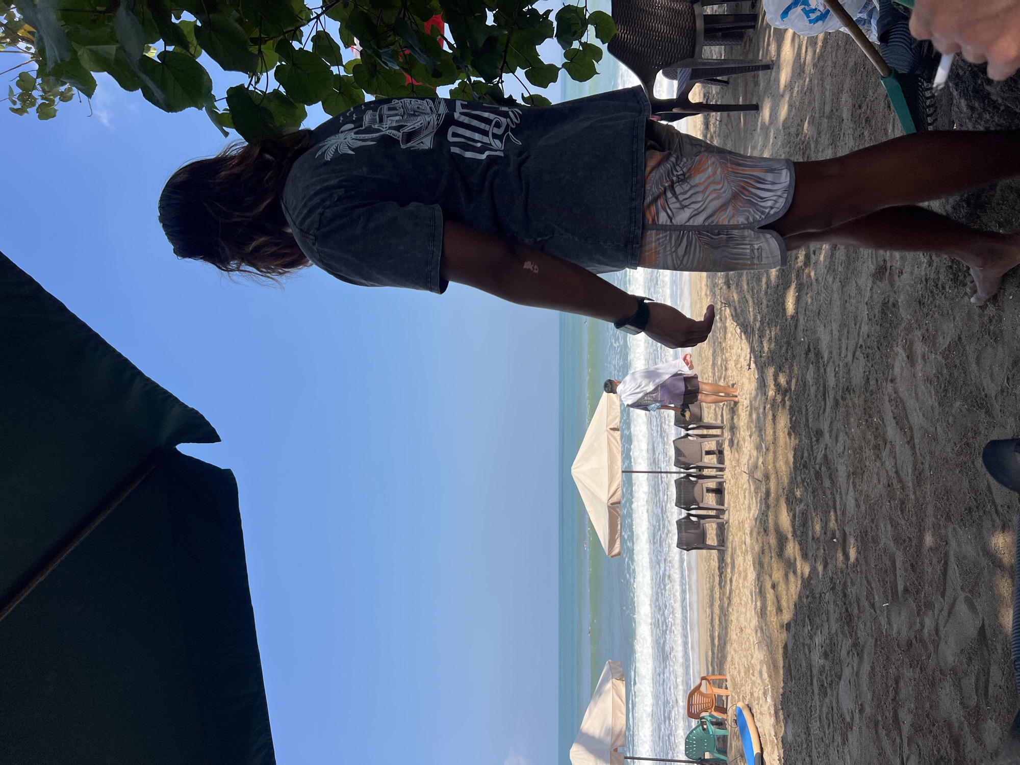 In Kuta, Indonesia, a person stands on a sandy beach, facing away from the camera and towards the ocean. The person is wearing a dark t-shirt with a graphic print on the back and patterned shorts. To the left, two beach umbrellas are set up, and several beach chairs are visible in front of them. The ocean is calm with gentle waves rolling onto the shore. In the distance, a few people can be seen in the water. The sky is clear and blue. The foreground of the image is dominated by sand and shadows cast by an overhead umbrella or tree. Another person is partially visible in the background, walking away from the water.