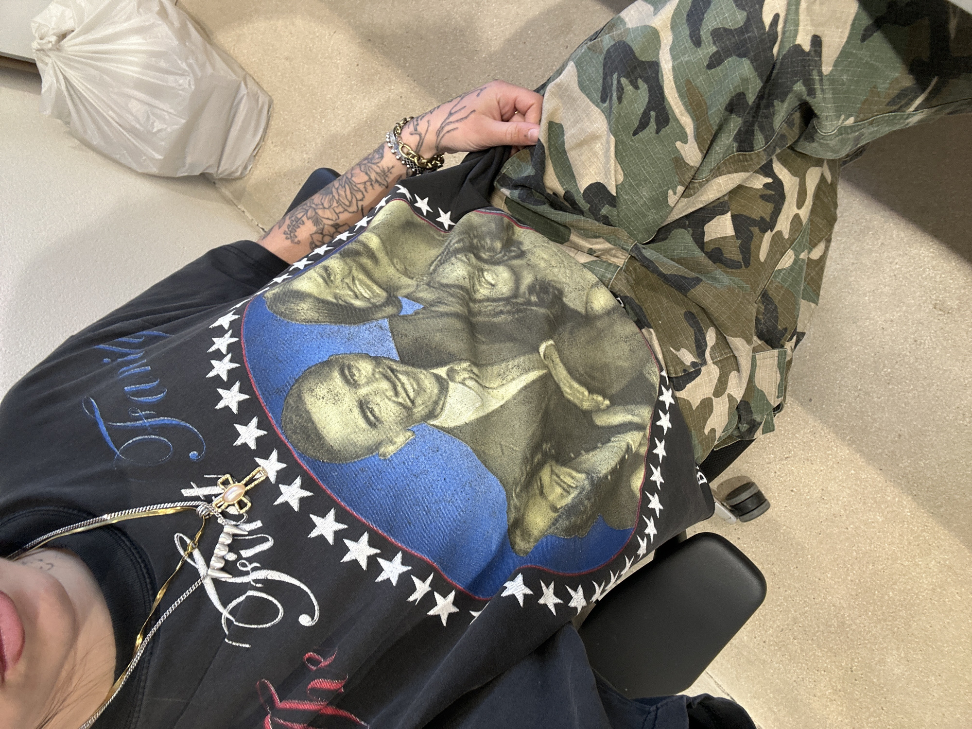 A person wearing camouflage pants and a black t-shirt with a blue circular graphic featuring a portrait surrounded by white stars sits in a black chair. The person has tattoos on their left arm and wears bracelets. A gold necklace hangs on the shirt. A white plastic bag is visible on the floor. The location is indeterminate and cannot be confirmed as Paris, France.