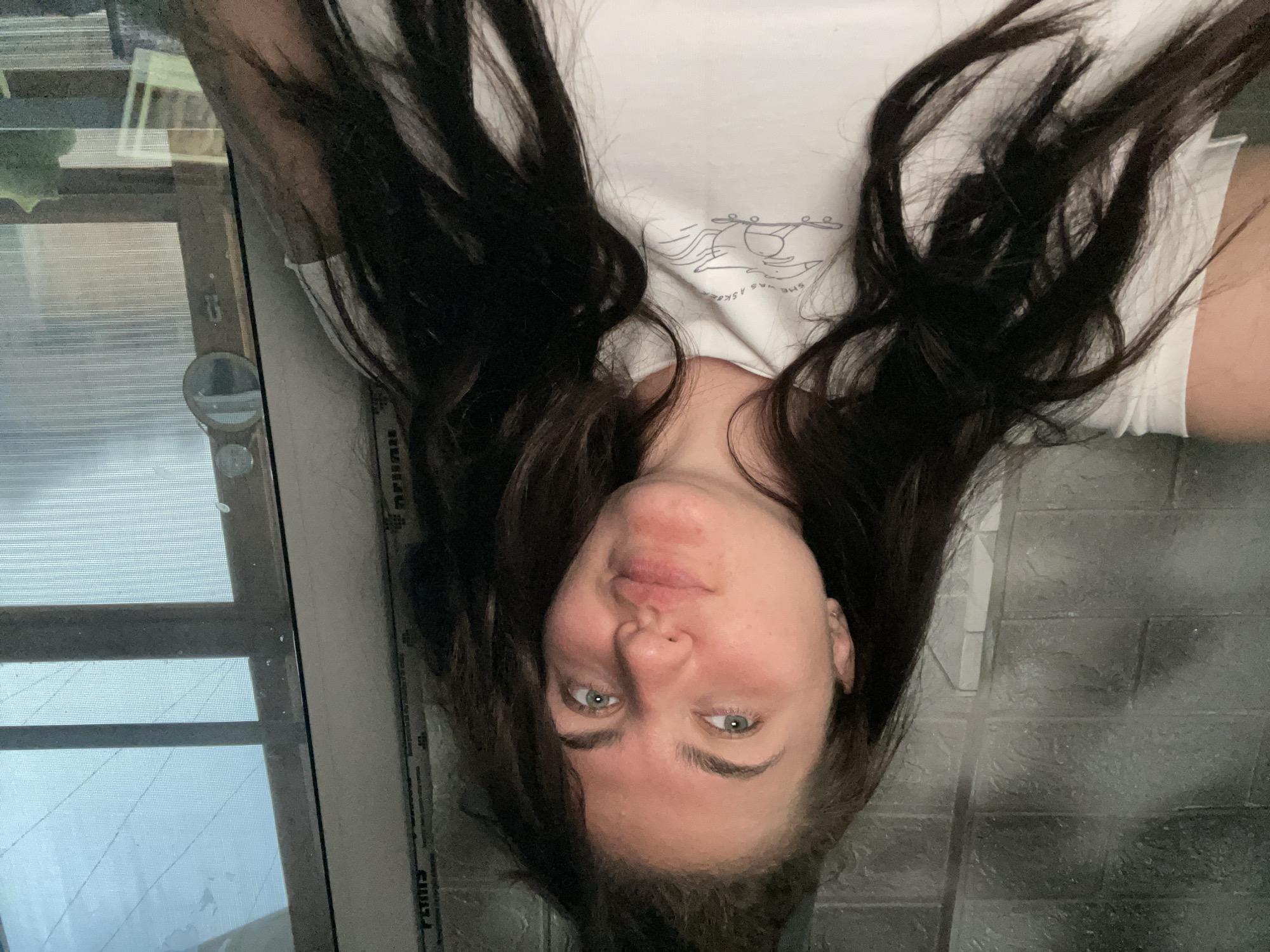 A woman with long, dark hair is lying down, facing the camera. She is wearing a white t-shirt with a graphic design. The background is blurry, but it appears to be a window with a screen. The location is likely in the city of Мосметаллоконструкция Онт, Russia.