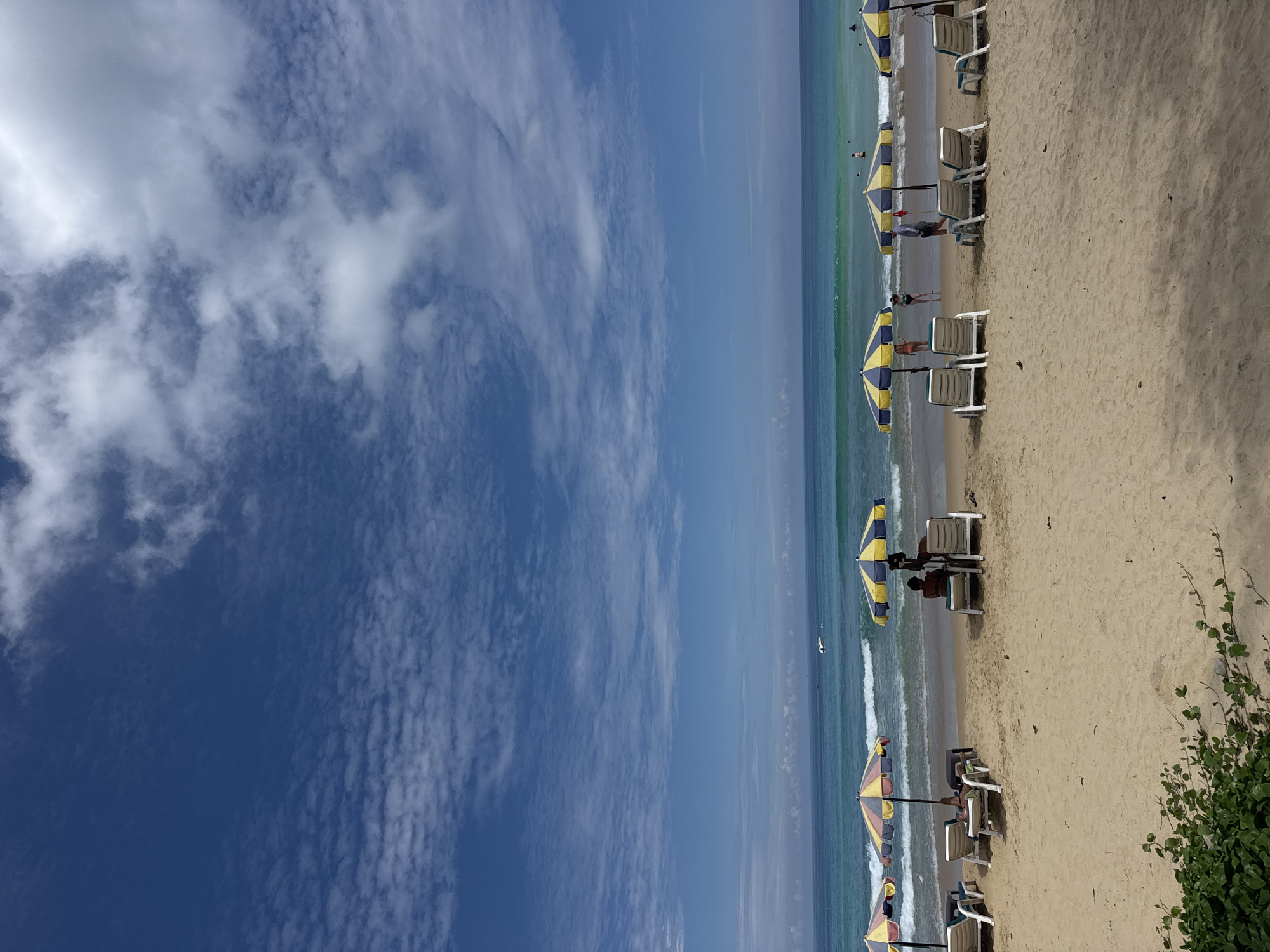The image depicts a beach scene under a bright blue sky with scattered clouds. The sand is light brown, and there are several beach umbrellas and lounge chairs set up. The ocean is a vibrant turquoise and blue, with moderate waves breaking on the shore. A few people are visible on the beach and in the water. In the distance, there is a small boat on the water. The overall atmosphere is one of a relaxed and sunny day at the beach.