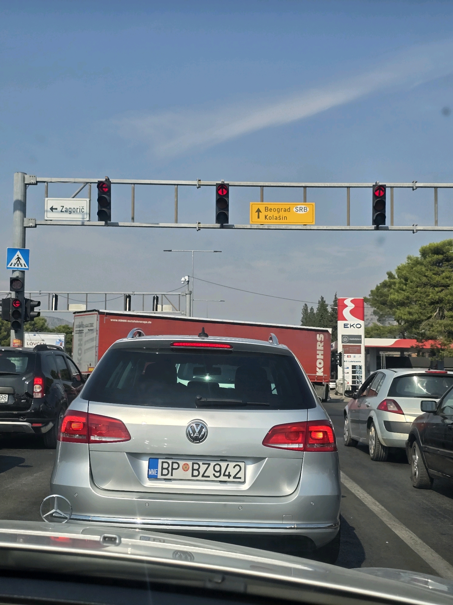 In Podgorica, Montenegro, a traffic jam occurs on a sunny day. Cars are stopped at a traffic light, with a silver Volkswagen station wagon in the foreground. Behind it, a red truck and other vehicles are visible. Overhead, traffic lights and road signs indicate directions to Zagorič, Beograd, SRB, and Kolašin. A pedestrian crossing sign and a security camera are also present. To the right, a red and white EKO gas station is situated with gas prices displayed. The sky is blue with some wispy clouds.