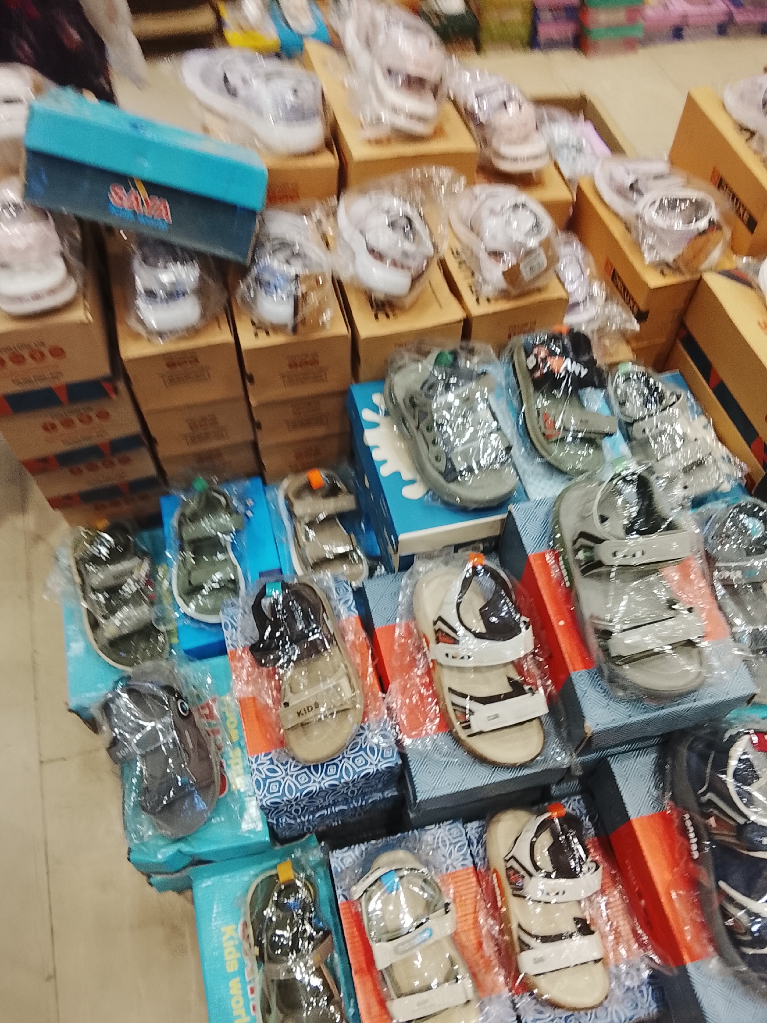 The image depicts a display of children's footwear in a shop, likely in Gulgasht Colony, Pakistan. The shoes are arranged on shelves and in boxes, mostly covered in plastic packaging. There are various styles of sandals and sneakers. The boxes are a mix of cardboard and colorful branded packaging. The scene is indoors, with an overhead perspective of the merchandise.