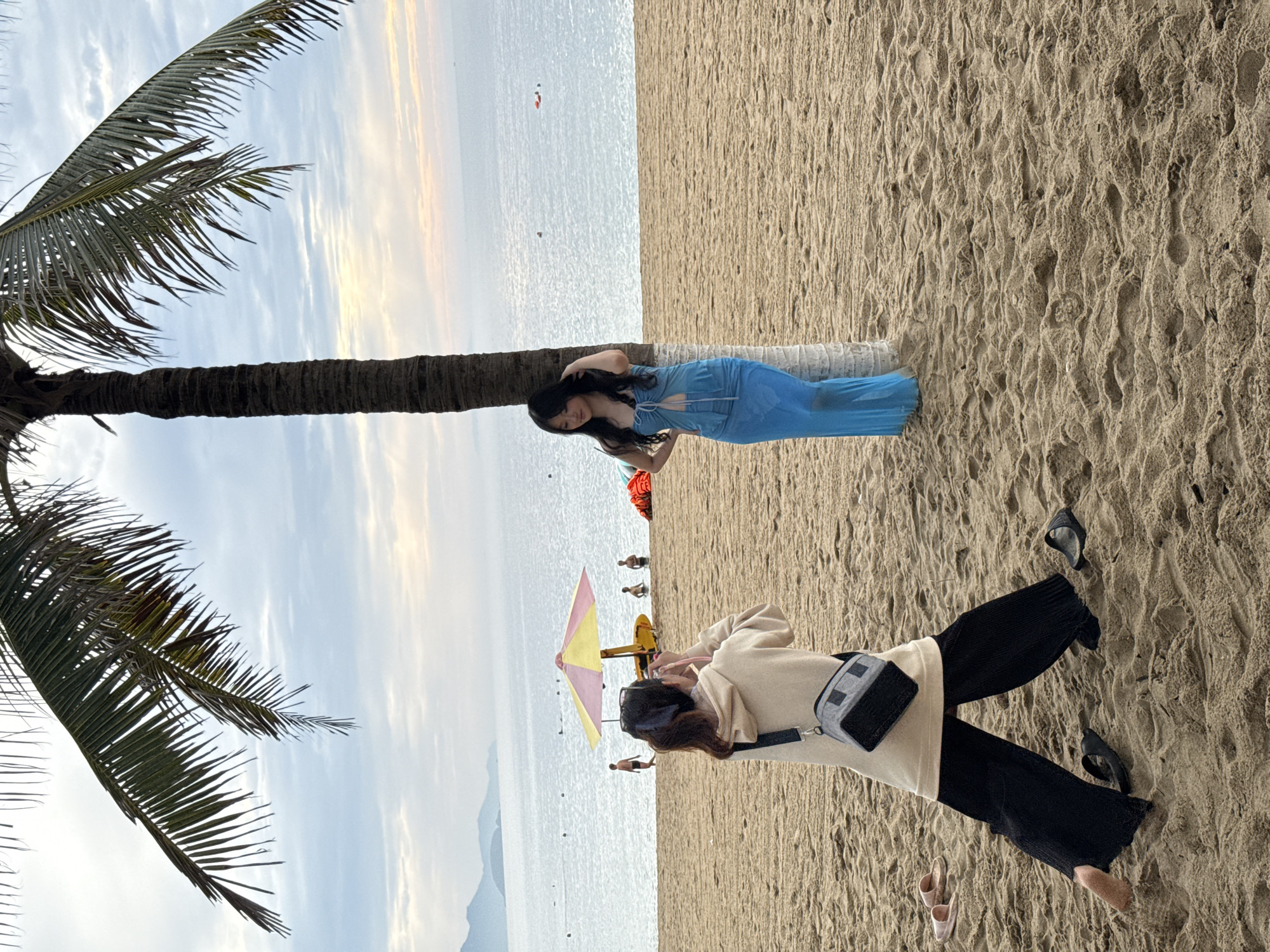 On a sandy beach in Nha Trang, Vietnam, a young woman in a blue dress poses near a palm tree. Another young woman in a beige hoodie and black pants is taking her photo. In the background, the ocean stretches to the horizon under a cloudy sky. A colorful umbrella and a yellow surf rescue float are visible near the water's edge, where several other people are present. The scene suggests a casual beach day with photography taking place.
