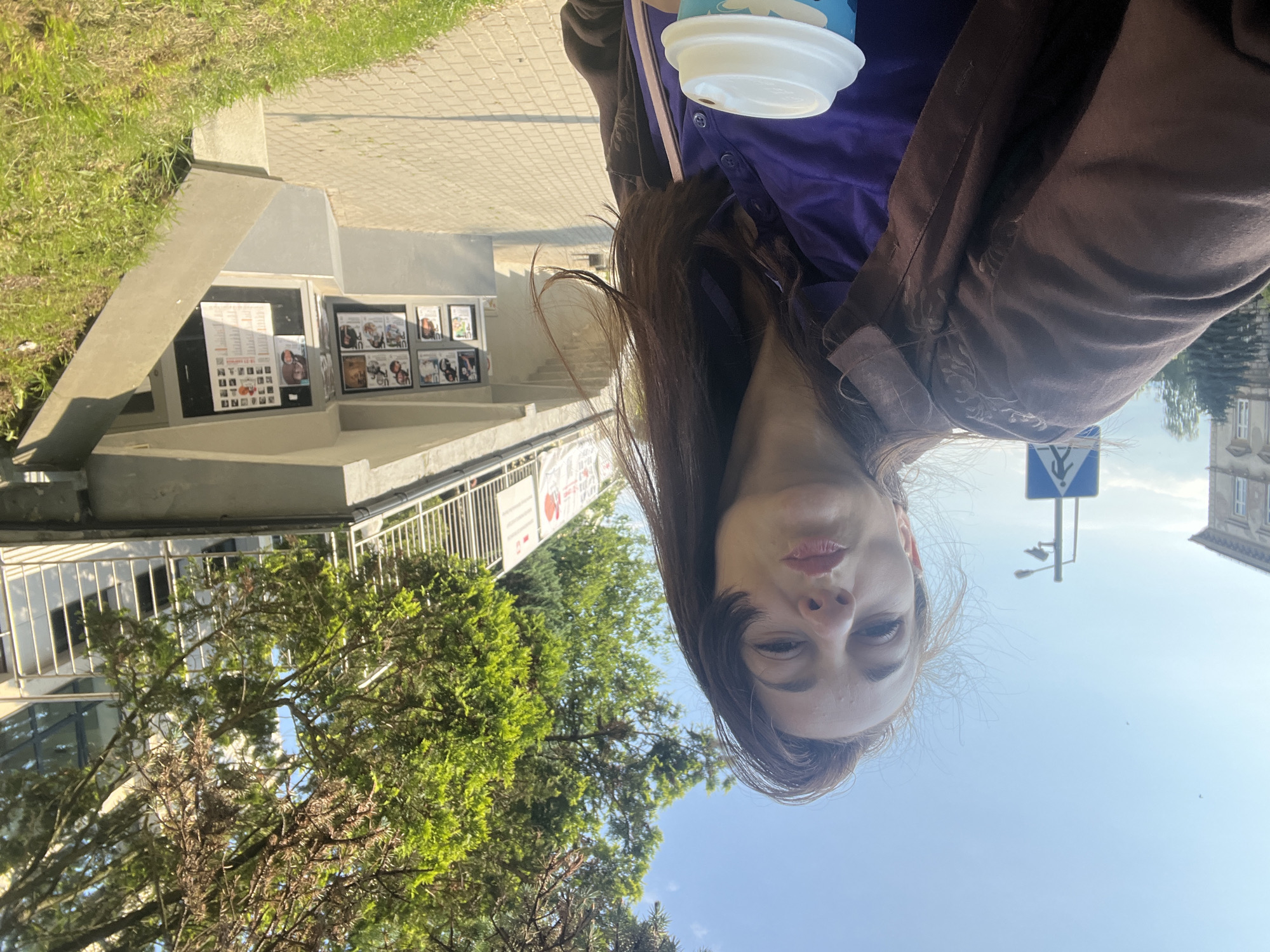 A woman with long brown hair is taking a selfie outdoors in front of a building with a ramp. She is wearing a brown jacket over a purple shirt and is holding a white coffee cup. She has her lips pursed as if kissing. In the background, there are trees, a sidewalk, a street sign, and posters displayed on the building. The sky is blue with a few birds flying.