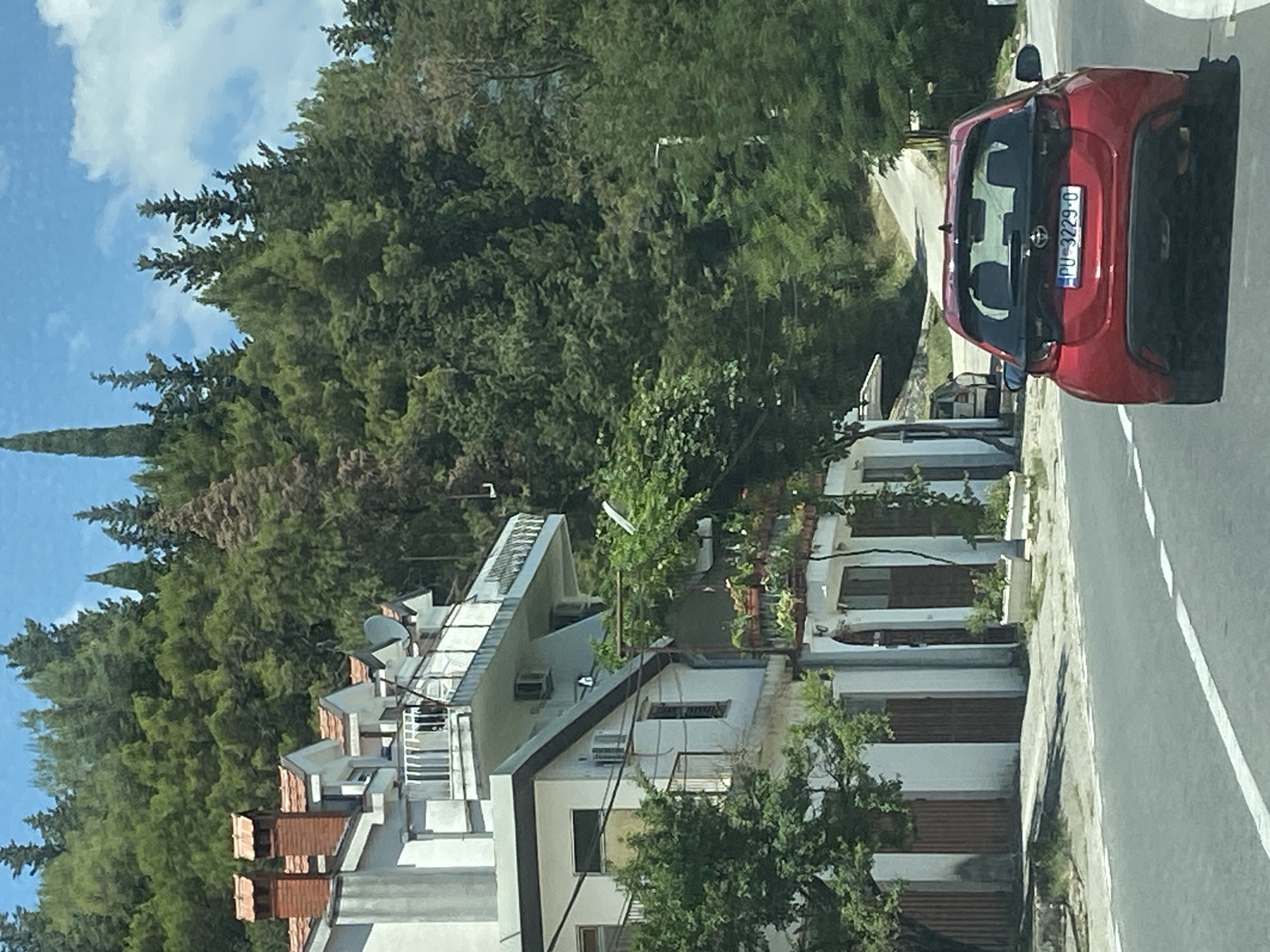 The image shows a red Toyota Yaris driving on a road in Sutorina, Montenegro, with a multi-story white building on the left and a dense forest on a hill behind it. The building features several balconies with white railings, red-tiled roofs, and chimneys. The forest is composed of numerous evergreen trees. The sky is blue with scattered white clouds. The car has a license plate that reads "PU 3229-O".