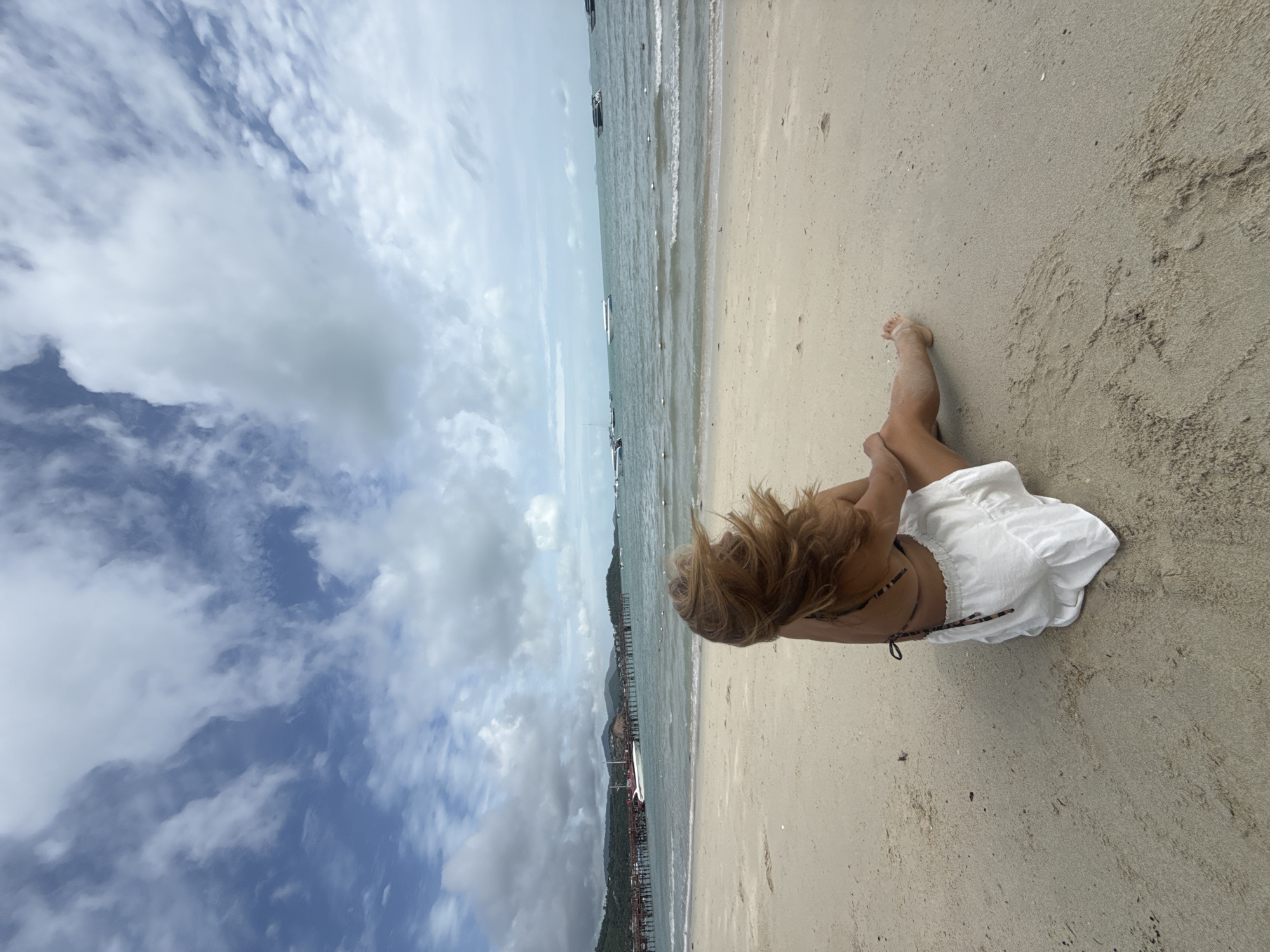 In the city of Ko Samui District, Thailand, a person with blonde hair sits on a sandy beach facing the ocean. The person is wearing a white skirt and a patterned bikini top. Their hair is blown by the wind. The ocean is calm with a few boats visible in the distance. A pier with buildings can be seen on the left side of the image. The sky is cloudy.
