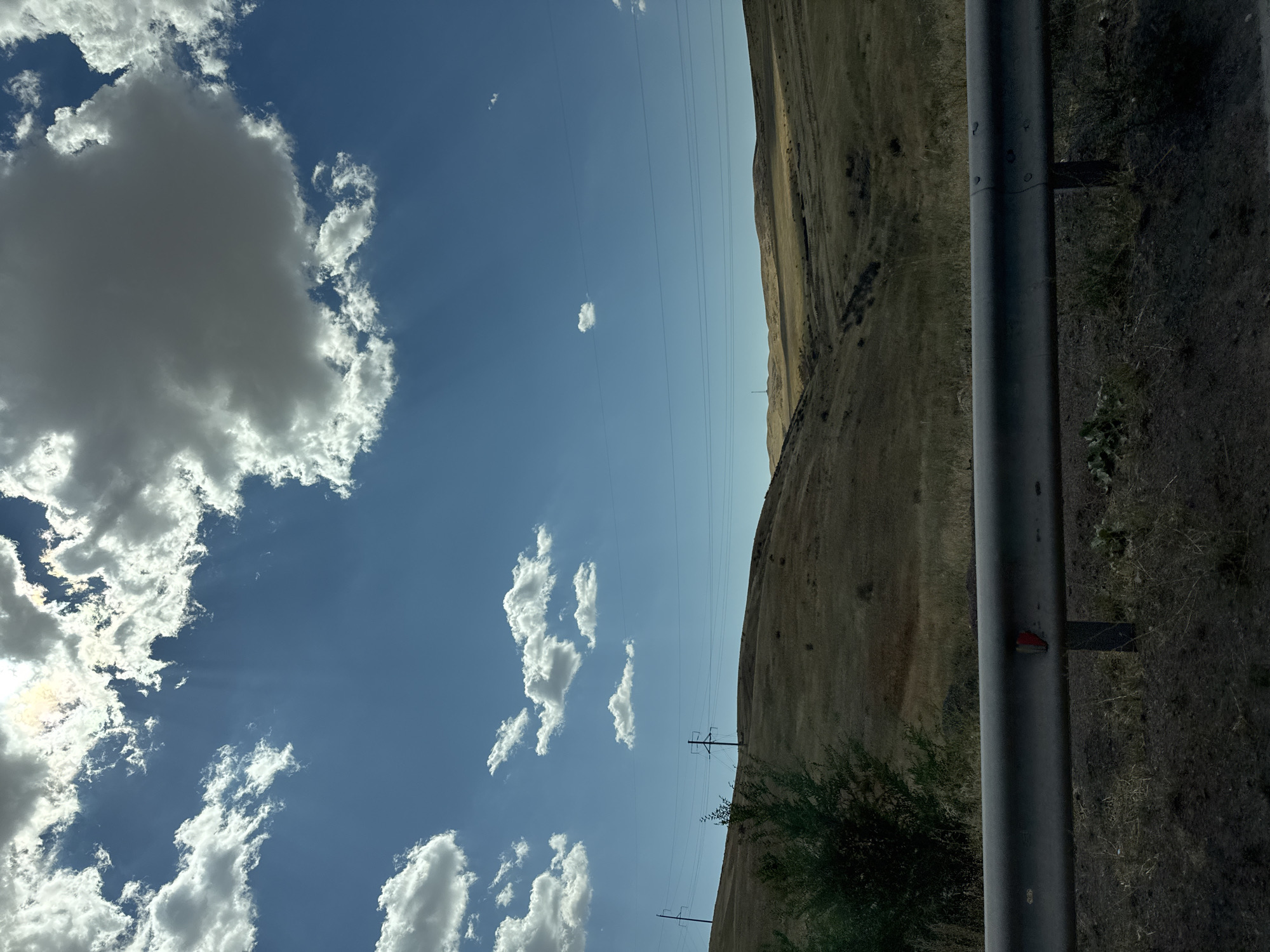 The image depicts a desolate, rolling landscape under a bright, cloudy sky. The terrain is covered in dry, golden-brown grass, suggesting a steppe or arid environment. The foreground features a silver metal guardrail, partially obscuring the ground, which is composed of dark soil and sparse vegetation.

In the mid-ground, a lone, sparse green bush is visible on the left. Power lines stretch across the scene, supported by utility poles that are sparsely distributed on the rolling hills. The sky is a vivid blue, punctuated by numerous white and grey clouds, some of which are illuminated by sunlight, creating beams of light that pierce through. One large, dark cloud dominates the upper right portion of the sky.

In the distance, more rolling hills are visible, their contours softened by the atmospheric perspective. The overall impression is one of isolation and natural, undeveloped scenery. The location is identified as the city of Arkharlinskii, Kazakhstan, which aligns with the described landscape characteristics of a steppe region. No people or specific structures other than utility poles are present.
