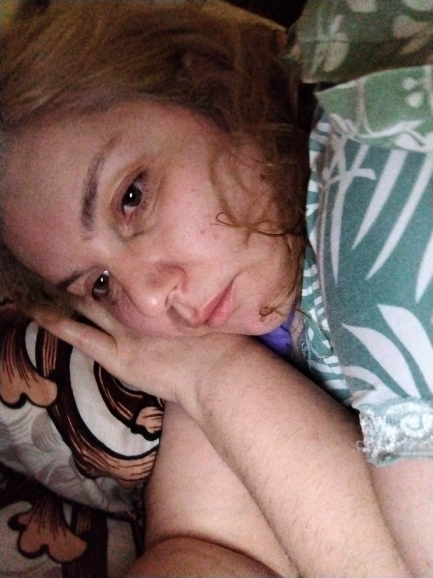 A person is lying down on patterned bedding with their head resting on their hand. The person has brown hair and brown eyes. They are wearing a garment with a green and white pattern. The background is blurred. The location is not discernible from the image.