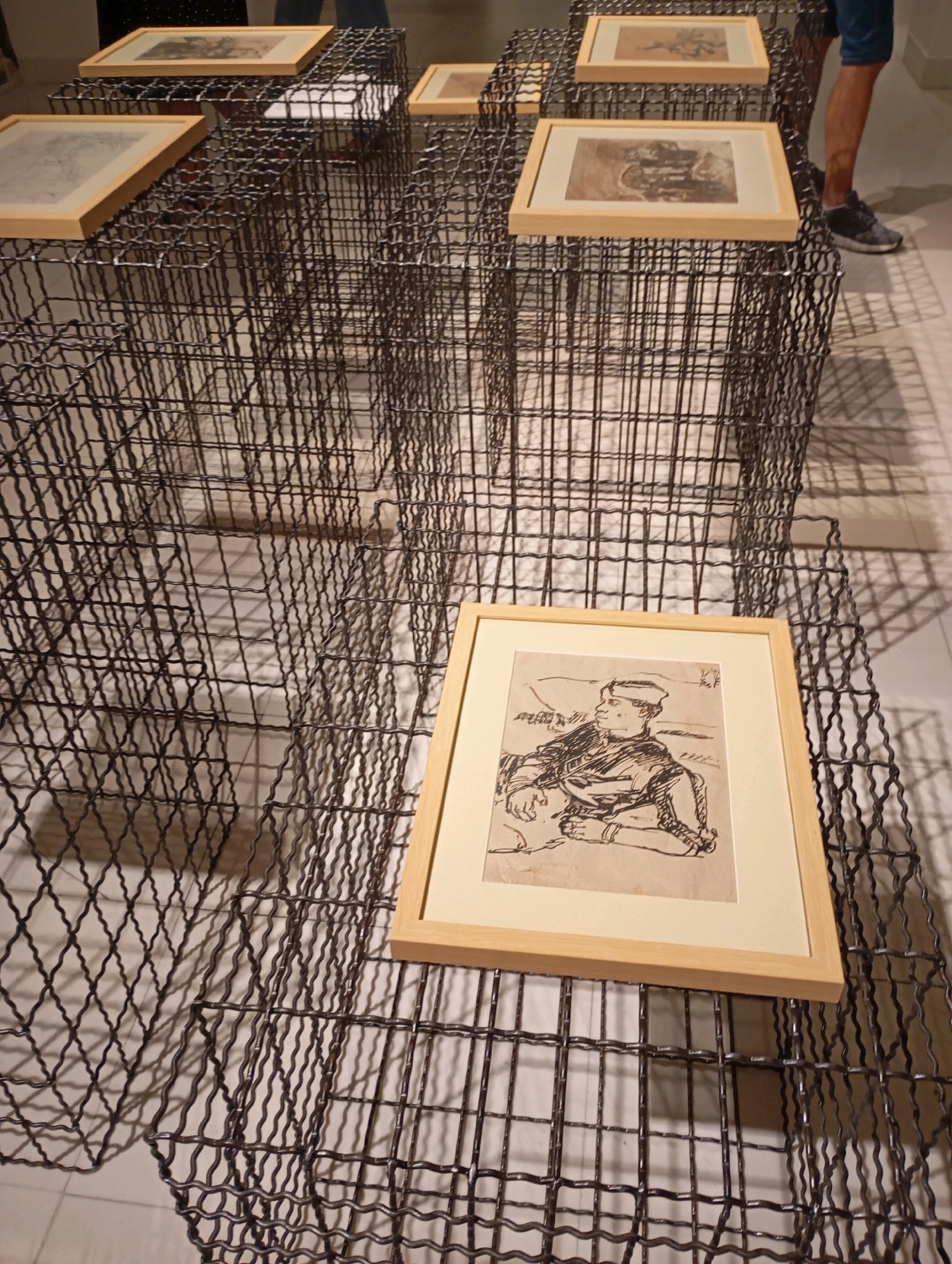 The image displays a gallery setting in Belgrade, Serbia, featuring framed artworks presented on wire mesh cubes. In the foreground, a framed sketch of a man in profile, rendered in black ink, rests on a wire cube. The sketch depicts the man in a seated position, wearing a hat and a jacket. The wire mesh structures, creating a grid-like display, cast shadows on the white floor, indicating ambient lighting. In the background, other framed pieces are visible on similar displays, some partially obscured by the foreground elements. A person's legs and feet, wearing shorts and athletic shoes, are visible on the right side of the frame, suggesting their presence as a visitor or staff member in the gallery. The overall scene conveys an artistic exhibition.