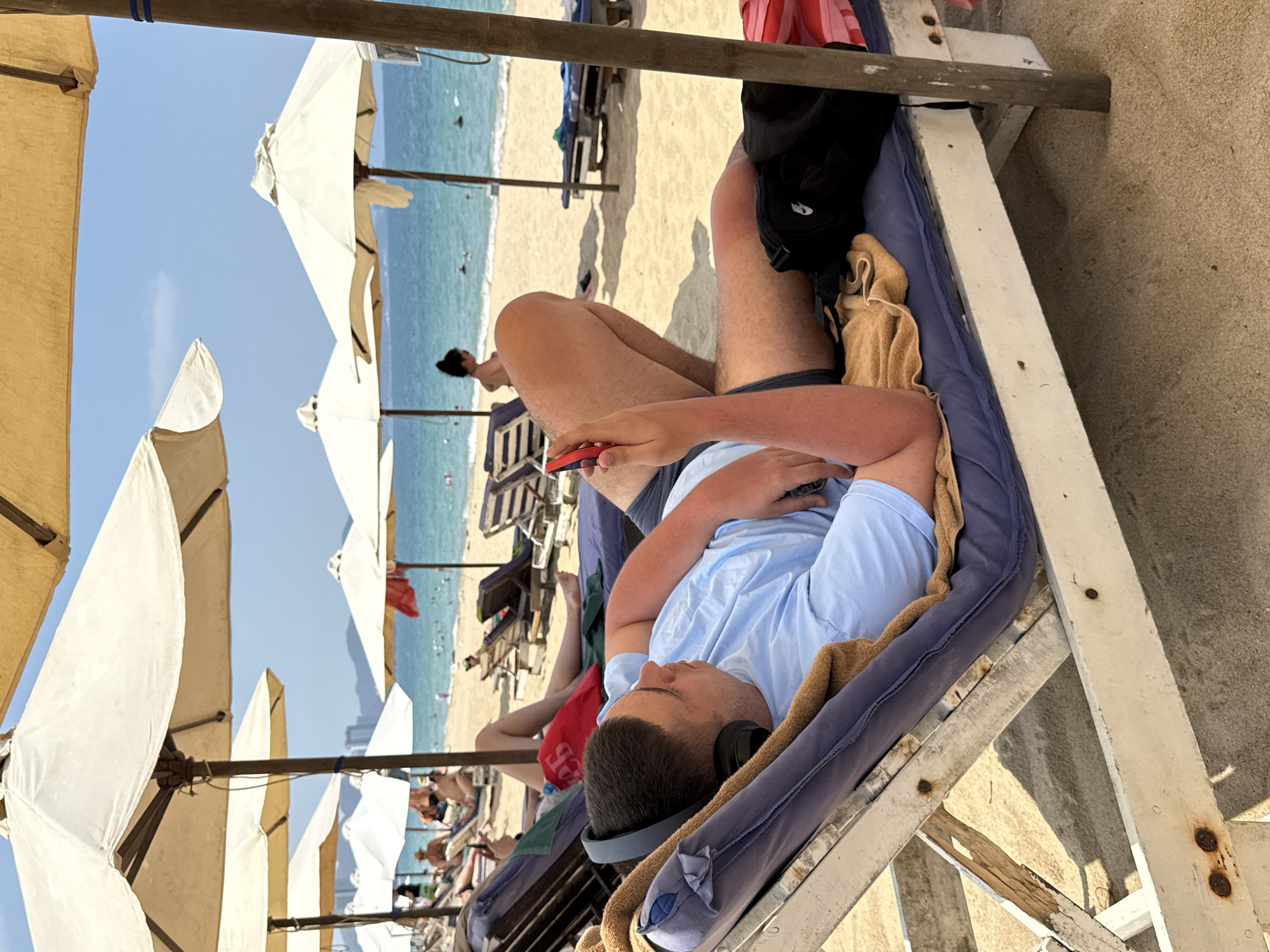 This image depicts a sunny beach scene in Nha Trang Ward, Vietnam, from a slightly elevated perspective. The main subject is a young man relaxing on a blue padded beach chair with a wooden frame. He is wearing a light blue t-shirt and dark shorts, with black headphones covering his ears and holding a smartphone with a red case. A brown towel is draped on the chair, and a black pouch is visible near his feet. His arms and legs show a reddish tint, indicative of sun exposure.
In the background, a sandy beach stretches towards the clear, turquoise ocean where several people can be seen swimming or wading. Numerous white and tan beach umbrellas are set up along the shore, providing shade for other beachgoers, some of whom are also visible relaxing on chairs. The sky is bright blue with minimal cloud cover, suggesting clear weather. The image conveys a leisurely atmosphere typical of a coastal resort. No visible text is discernible within the frame.