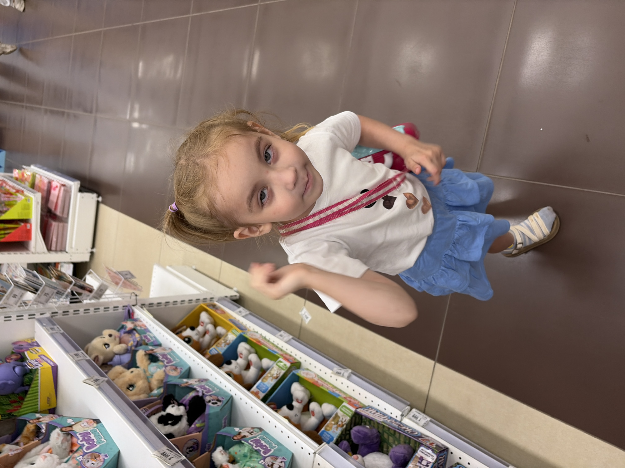 A young Caucasian girl with blonde hair in a ponytail stands in an aisle of a toy store. She is wearing a white t-shirt with heart appliques, a blue skirt, and sandals. A pink and white striped strap is visible across her chest. The girl is looking at the camera with a slightly squinted expression. Behind her, shelves are stocked with various plush toys, some in their original packaging. The floor is tiled with dark brown and beige sections. The image appears to have been taken in a retail environment, possibly a large department store or specialized toy shop. There is no specific indication of the location being in Tambov, Russia, beyond the provided prompt.