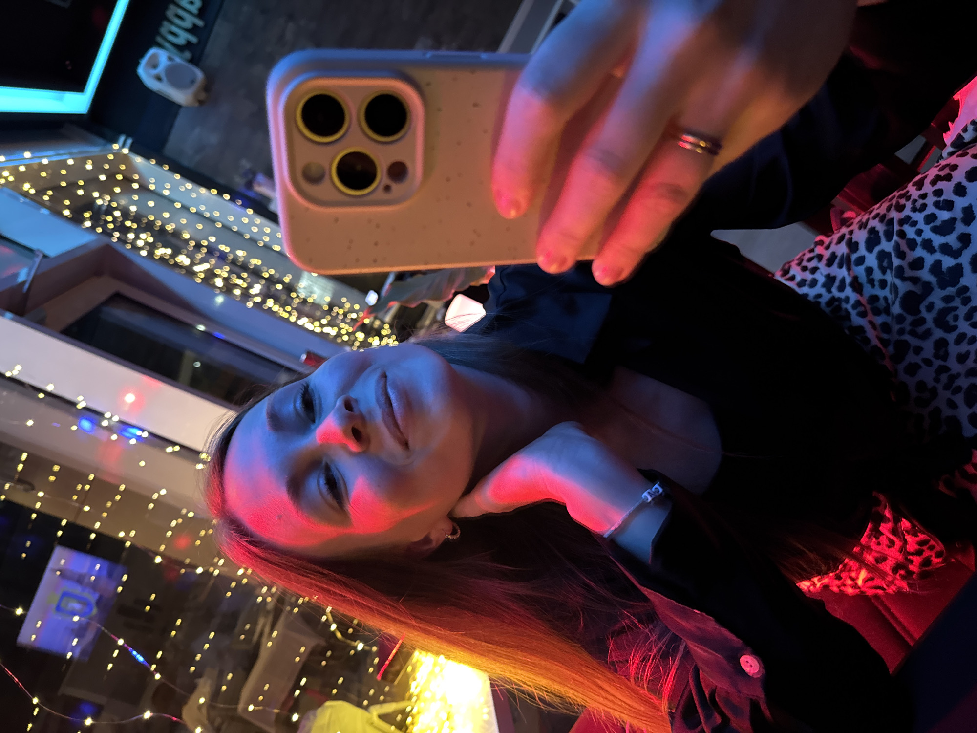A person with fair skin and light brown hair is taking a selfie with an iPhone. The phone has a light grey case and three camera lenses are visible. The person is wearing a black shirt, a bracelet, and a ring. The background features a window with decorative string lights, suggesting an urban setting. Based on the context of Budva, Montenegro, this scene likely takes place in a bar or restaurant with outdoor seating near the old town, and "gabbb" text is visible on the window. There is a chair in the background of the shot with an animal print pattern. The lighting is mixed with blue and red hues, which is not the correct hue, however, given the lights it does correctly highlight the overall composition.