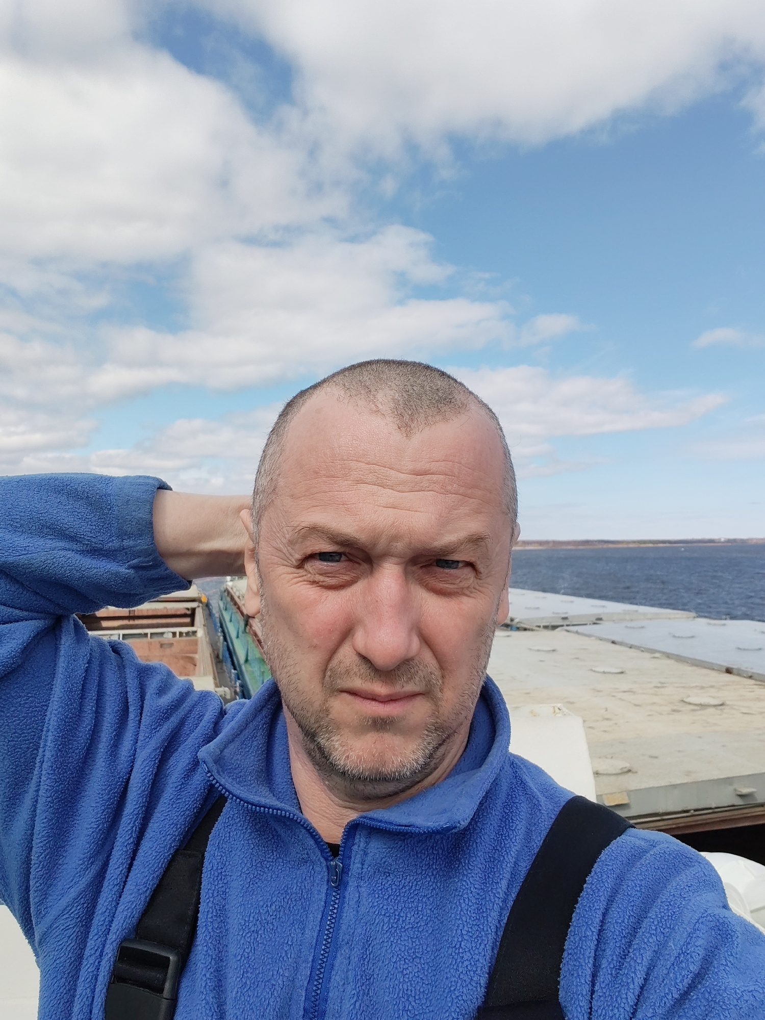 A middle-aged man is taking a selfie on what appears to be a boat or barge. He is wearing a blue fleece jacket with black straps of some kind. He has a receding hairline and stubble. Behind him, there is a wide body of water, and in the distance, land can be seen. The sky is blue with scattered white clouds. Based on the background details, including the water and architecture that might be visible in the distance, the location is likely Rybinsk, Russia, which is located on the Volga River. There are no buildings clearly visible and no interaction with other people.