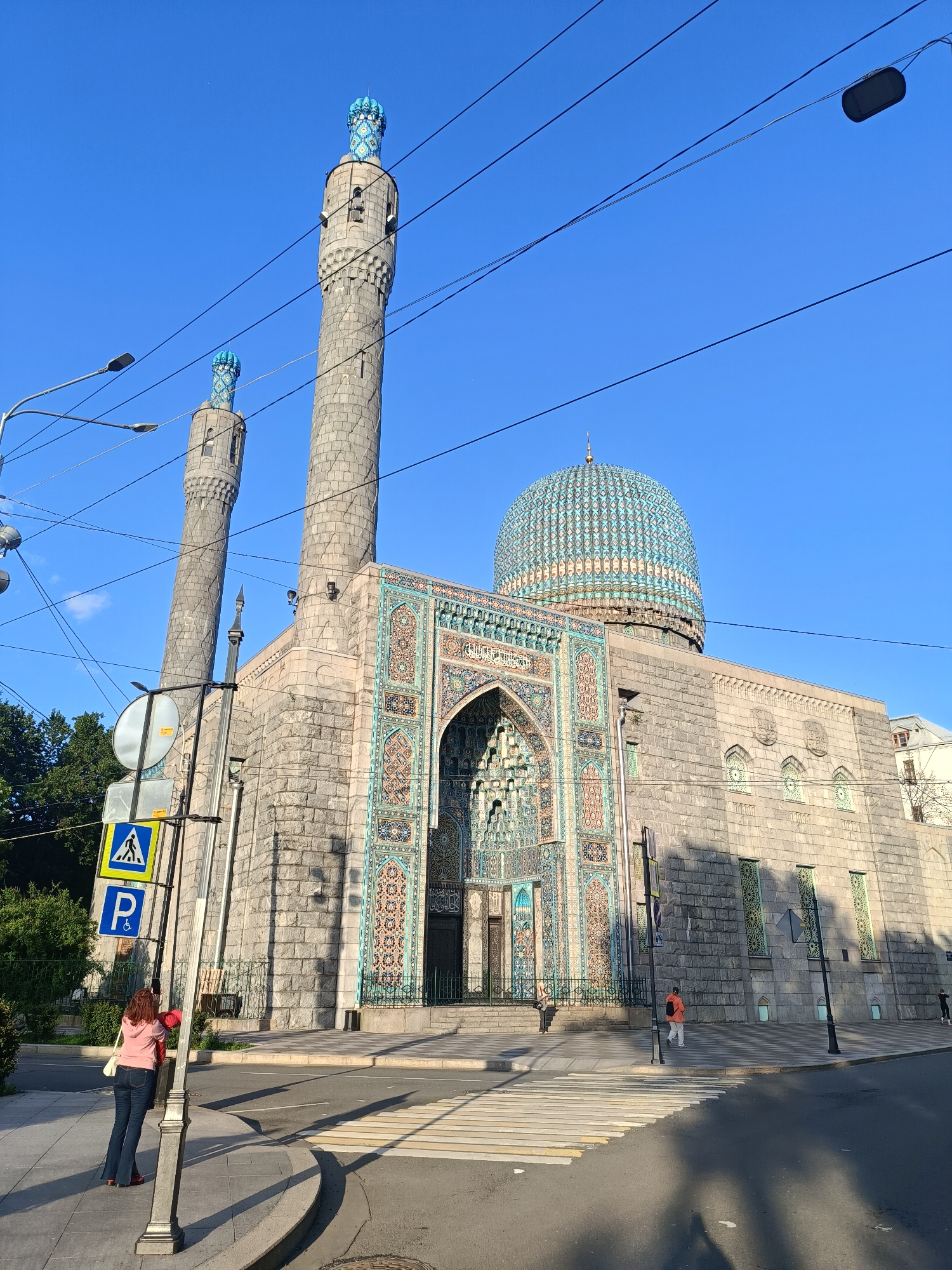 The image shows the Saint Petersburg Mosque, a prominent landmark in Saint Petersburg, Russia. The mosque is an impressive structure with grey stone walls, two tall minarets topped with blue and gold patterned domes, and a large, ornate blue dome. The entrance to the mosque is decorated with intricate mosaics in shades of blue, turquoise, and orange, featuring geometric patterns and Arabic calligraphy.
In the foreground, a pedestrian crossing is visible on the street. On the left sidewalk, a woman in a pink hooded jacket and blue jeans stands near a lamppost with traffic signs, including a pedestrian crossing sign and a parking sign. She is holding a mobile phone. Further down the sidewalk, another person wearing an orange jacket and white pants walks away from the camera. The building is bathed in sunlight, casting long shadows across the street and sidewalk. The sky is clear and blue. Trees are visible behind the mosque on the left side of the image. The architectural style of the mosque is a blend of Moorish and Byzantine influences.