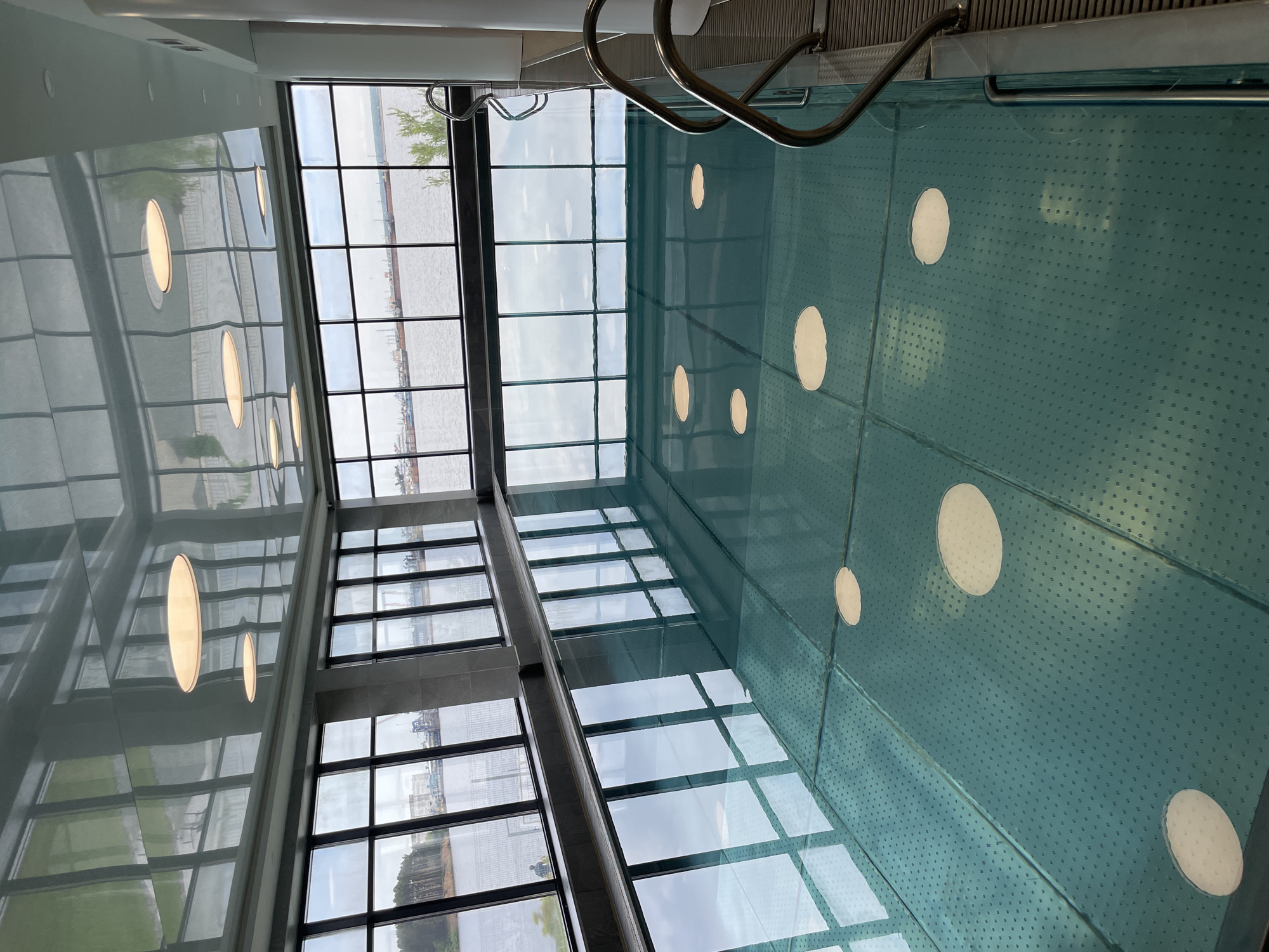 This image captures an indoor swimming pool area, viewed from a high angle looking down into the pool and across towards large windows. The swimming pool features clear, light blue water with a textured pattern of small dots on its bottom or side surfaces. Several large, circular, illuminated fixtures are embedded within the pool itself, casting a bright glow. Metal handrails for entering and exiting the water are visible in the upper right portion of the frame.
To the left of the pool, expansive floor-to-ceiling windows reveal an outdoor scene. Through these windows, a wide body of water, likely a river or bay, is visible, extending towards a distant shore with what appear to be industrial or harbor structures, including buildings and potentially cranes. Some greenery is also discernible. The sky outside is bright but appears cloudy or overcast, indicating daytime.
A highly reflective surface, possibly a ceiling or an adjacent wall, runs along the left side of the image, showing strong, distorted reflections of the windows, the outdoor environment, and elongated oval-shaped light fixtures. No people are present in the scene, and no visible text can be identified. The overall impression is of a modern, well-lit aquatic facility offering scenic views.
