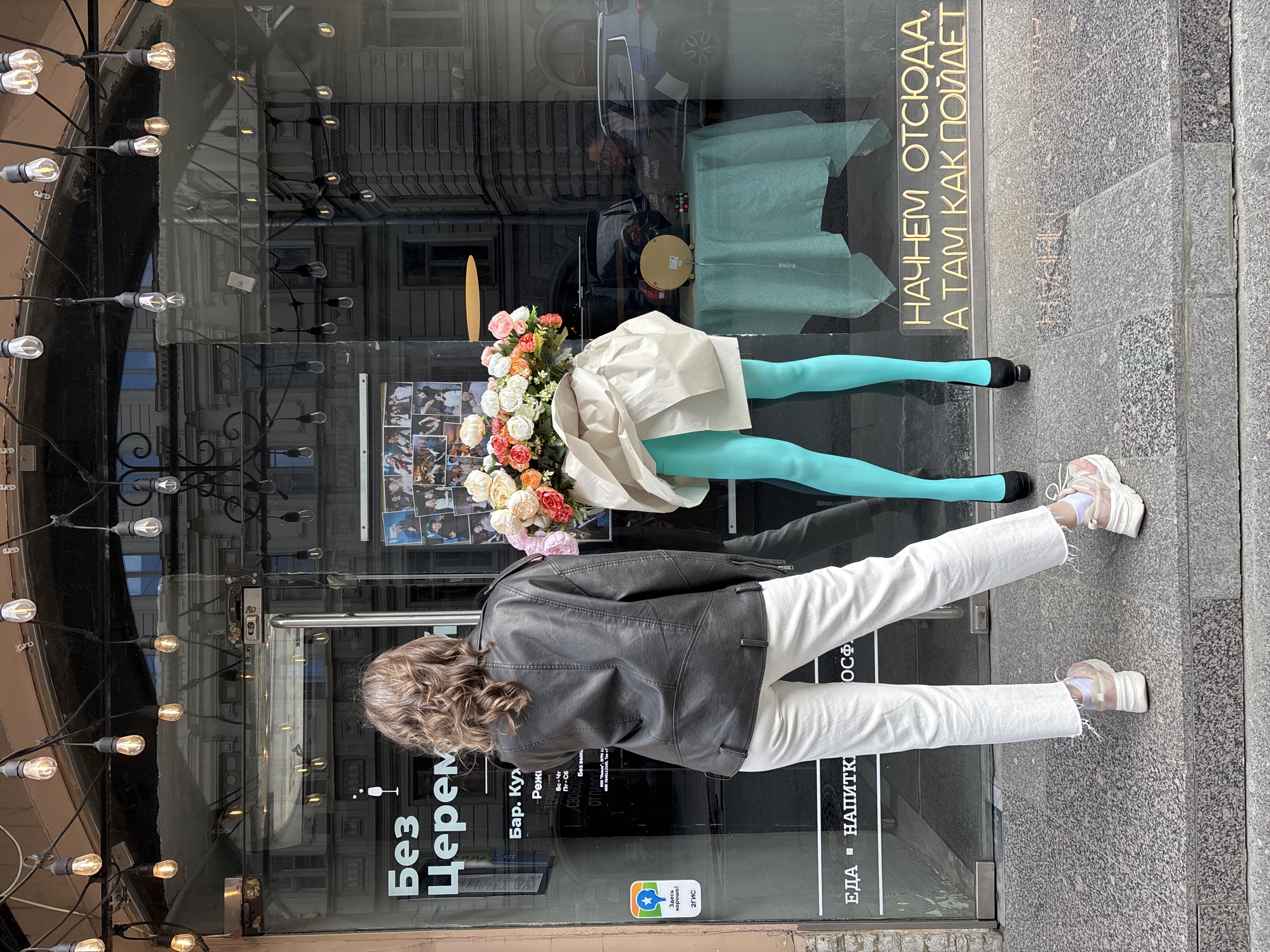 A woman in a black leather jacket and white pants stands in front of a business entrance. The business appears to be located in Saint Petersburg, Russia, as the business logo uses Cyrillic script. The business logo partially visible on the glass door reads "Без Церем". The business also features a 2GIS logo. A mannequin is displayed in the business window. The mannequin's head is replaced with an arrangement of flowers, and it wears blue tights and a short white garment. The window reflects the surrounding cityscape. A string of lights hangs next to the glass door. The ground is paved with gray stone slabs.