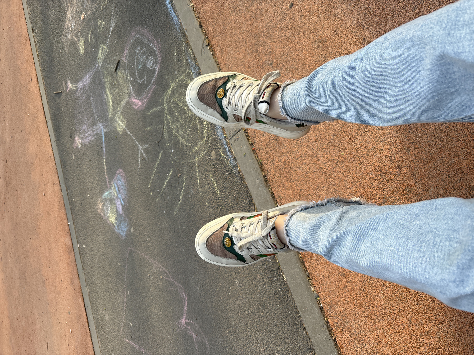 The image shows a person's legs and feet from an overhead perspective. The person is wearing light blue denim jeans and a pair of designer sneakers that appear to be Gucci. The shoes are off-white with brown and green accents.
The person is standing on a surface divided into three sections: one side is a reddish-brown textured material, a central section of asphalt with chalk drawings, and a thin strip that is a different shade of gray.
The location is potentially Saint Petersburg, Russia, considering the urban setting and architectural style, however, more evidence is needed to confirm this.