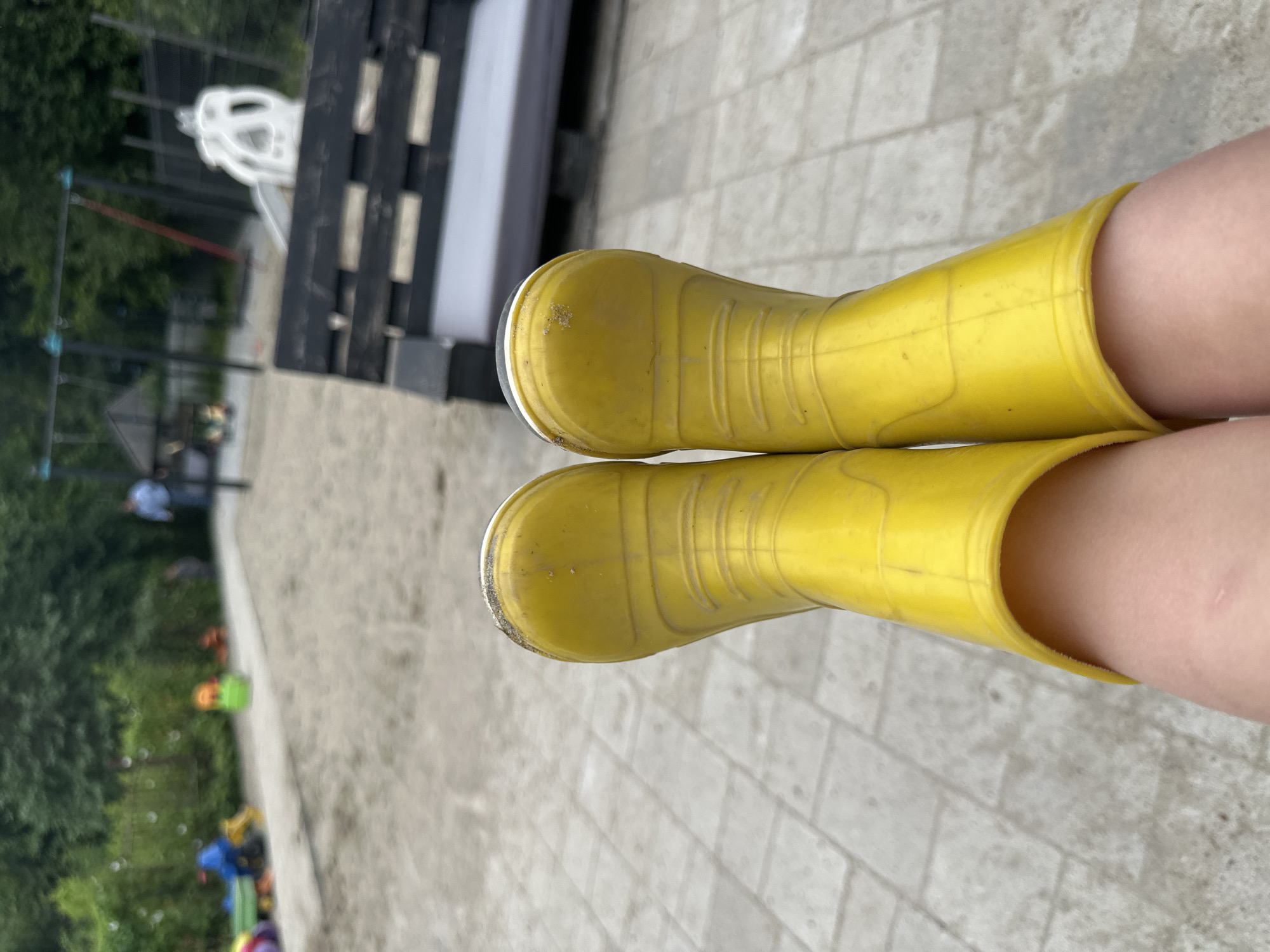 The image shows a close-up perspective of a person's legs wearing bright yellow rubber boots. The boots appear to be slightly dirty. The background suggests an outdoor setting, possibly a playground or park area with sand and paved ground. In the distance, playground equipment such as swings and a slide are visible, along with some greenery from trees and bushes. There are a few blurred figures of people in the background, including one person wearing a blue shirt and another in a blue jacket. A black bench made of wooden slats is in the mid-ground. The overall impression is that of a casual, outdoor environment suitable for children's play. The location of Malinovka, Russia, cannot be determined from the visual information provided in the image.