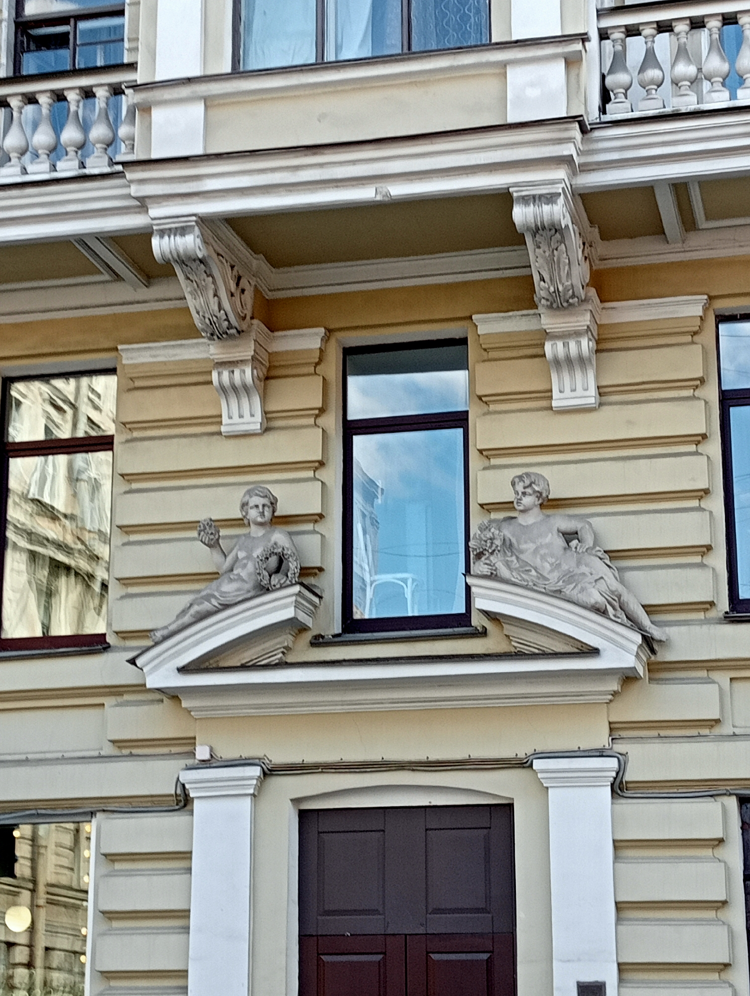 This is a photograph of the facade of a building in Saint Petersburg, Russia, featuring classical architectural elements. The building is painted a pale yellow, with white decorative moldings and columns.
Two stucco sculptures of putti, or cherubs, are prominently displayed on the facade, positioned on either side of a central window. These figures are reclining and appear to be holding wreaths. The putto on the left is holding a bundle of what may be fruit or flowers in its left hand and a wreath in its right. The putto on the right is holding a wreath of flowers. The sculptures are light-colored and weathered.
Above the sculptures, there is a horizontal balcony with a white balustrade, consisting of turned balusters and a handrail. Decorative brackets with carved details support the structure above the balcony.
The building has several windows with dark frames. One large window to the left of the sculptures reflects the surrounding buildings, showing architectural details and windows of another structure. A central, tall window above the sculptures has a blue tint, likely due to the glass, and also reflects the sky.
Below the sculptures, a large, dark brown double door is visible, set within an arched entryway flanked by white columns. The lower section of the building exhibits a rusticated stone pattern.
The overall impression is of an ornate and historical building, characteristic of Saint Petersburg's architectural style. The lighting suggests it is daytime.