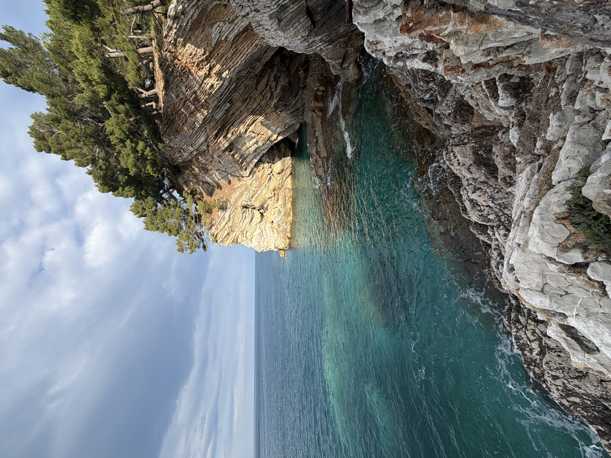 The image shows a rocky coastline near Petrovac centar, Montenegro. The scene is dominated by layered rock formations along the coast, with trees clinging to the cliff tops. The water is clear and turquoise, contrasting with the grey and brown tones of the rocks. The sky is partly cloudy. There are no people visible.