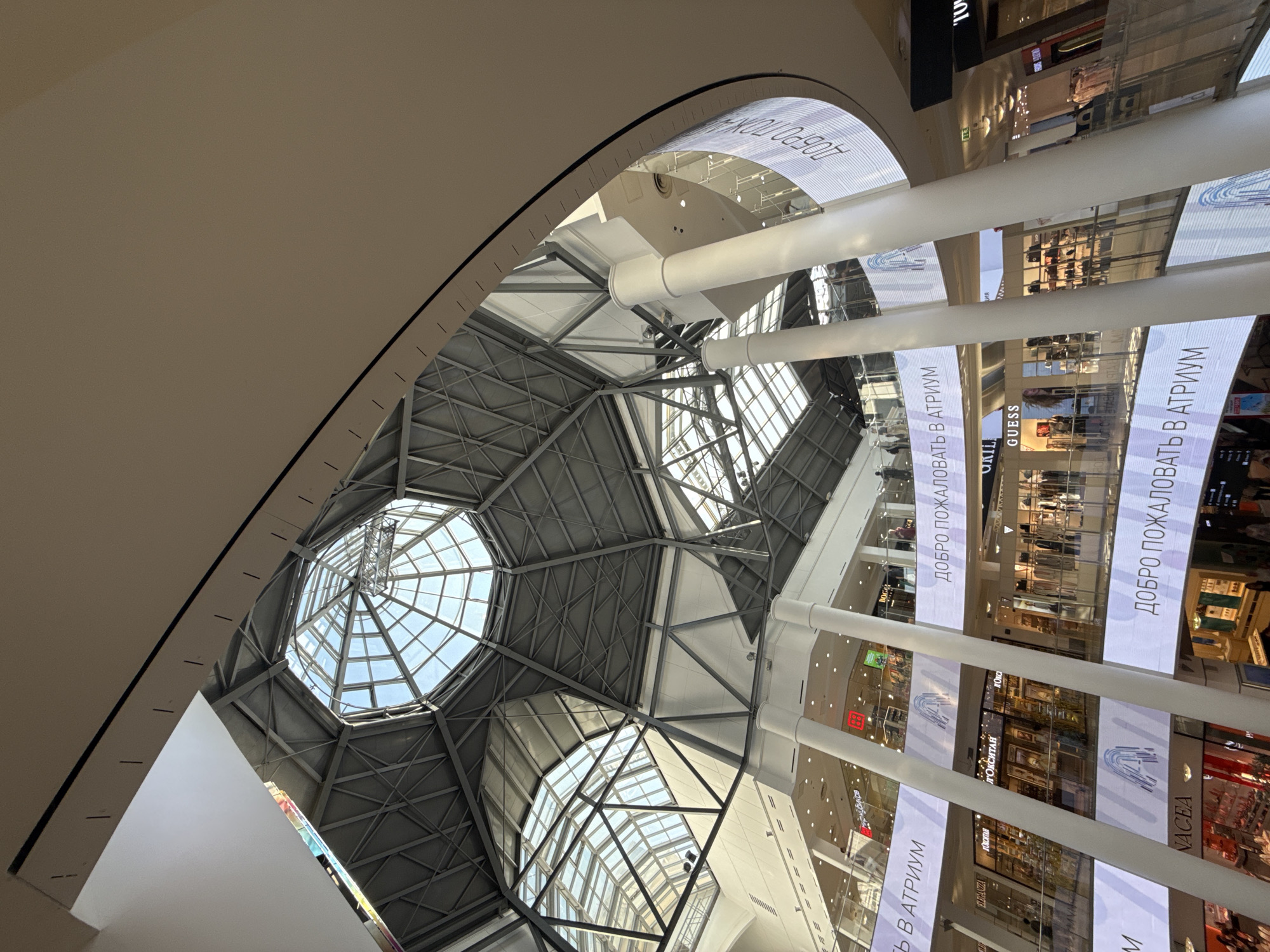 The image captures an interior perspective looking upwards towards the multi-level atrium of a modern shopping mall in Moscow, Russia. A striking architectural feature is the large, domed skylight constructed with a web of metal beams and glass panels that filter natural light. The mall consists of multiple floors, evident from the receding tiers of retail spaces, delineated by pillars. Prominently displayed on digital screens attached to the pillars is the phrase "ДОБРО ПОЖАЛОВАТЬ В АТРИУМ", which translates to "Welcome to the Atrium". Brand names such as "GUESS" and "Л'ОКСИТАН" indicate the presence of recognizable retail establishments.
