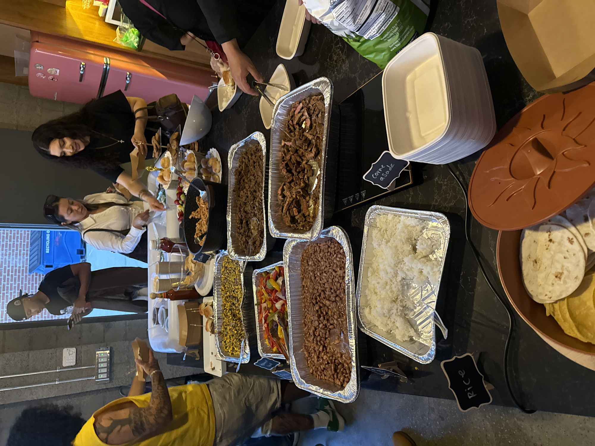 The image shows a food buffet setup, likely for a gathering. The buffet includes multiple aluminum trays containing various foods such as shredded beef, carne asada, beans, rice, sauteed peppers and onions, and corn. Next to the food trays are stacks of tortillas and bowls. People are shown serving themselves from the buffet. The location is Culver City, United States of America based on a recycling bin in the background.