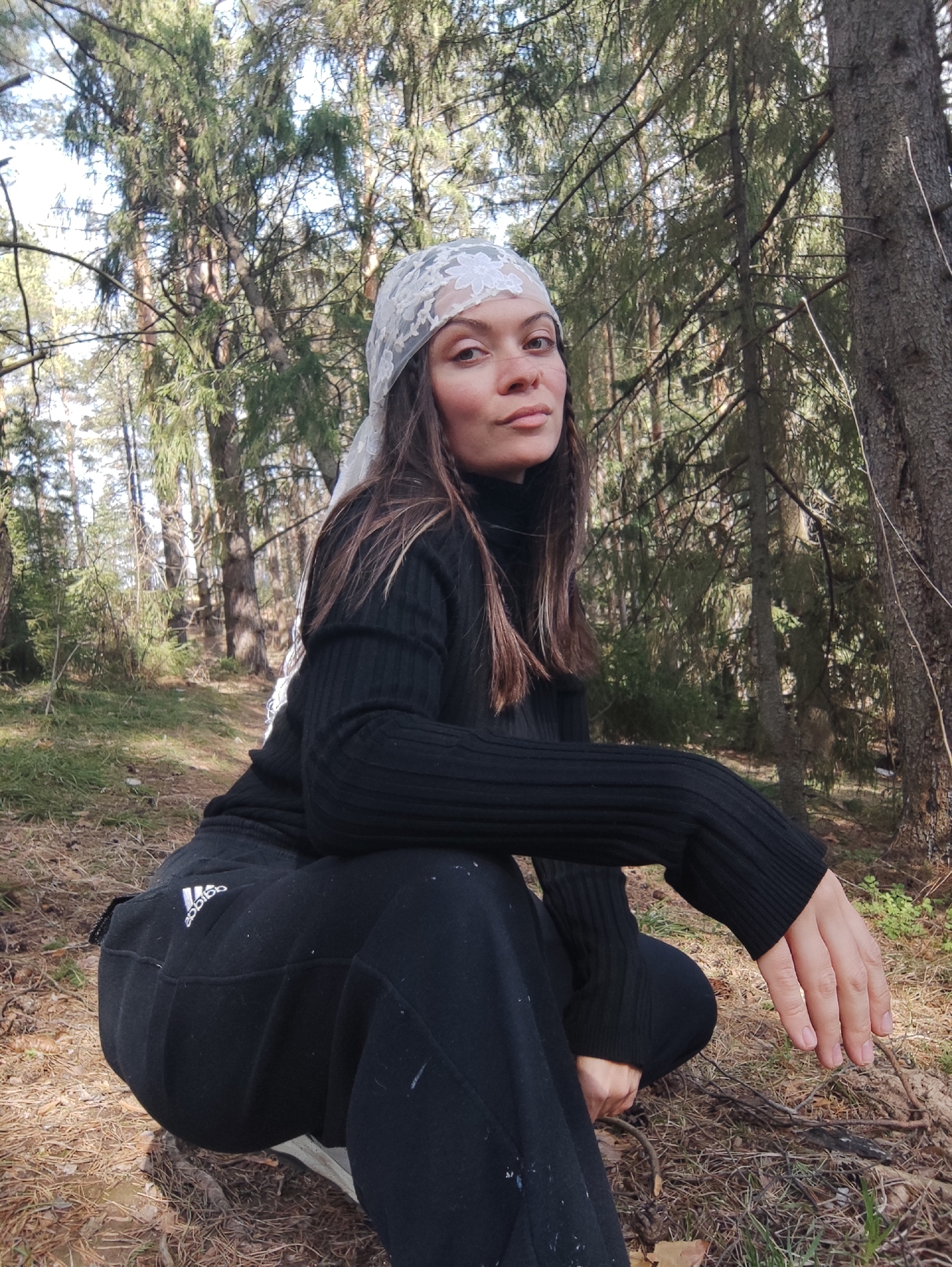 The image shows a woman crouching in a forest. She is wearing a black turtleneck sweater, black sweatpants with the Adidas logo, and a white lace headscarf. Her hair is long and brown. The forest setting includes tall trees and a forest floor covered with leaves and pine needles. The location is in Фоки, Russia.