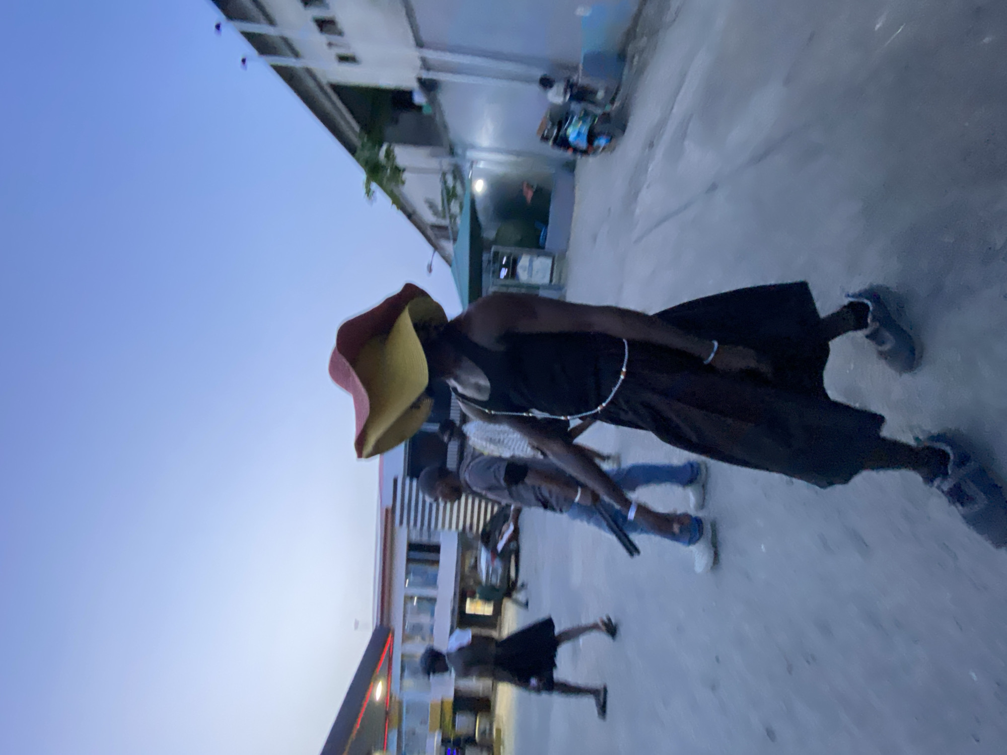 A street scene in Ikorodu, Nigeria. A person wearing a red and yellow hat, a black tank top, and a long black skirt or dress is walking. Two men are standing nearby. A fourth person is walking toward a building with red accents. A building with a green awning and two people sitting on a bench is behind the person with the hat. The sky is pale blue.