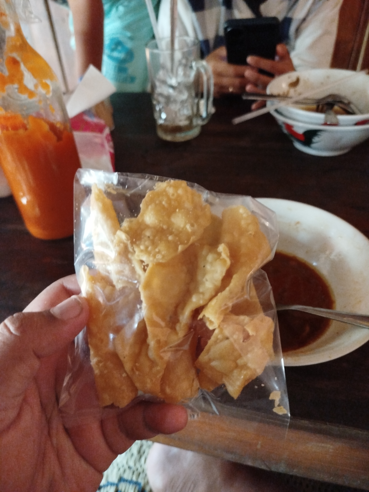 In Pojok, Indonesia, a person's hand is holding a clear plastic bag filled with fried snacks. The snacks appear to be golden-brown and irregularly shaped. To the right of the bag, a white bowl contains a dark sauce, with a spoon resting inside. In the background, a glass of ice is visible, along with a person holding a dark-colored smartphone. A bottle of orange sauce is partially visible on the left side of the image. The scene is set on a wooden table.