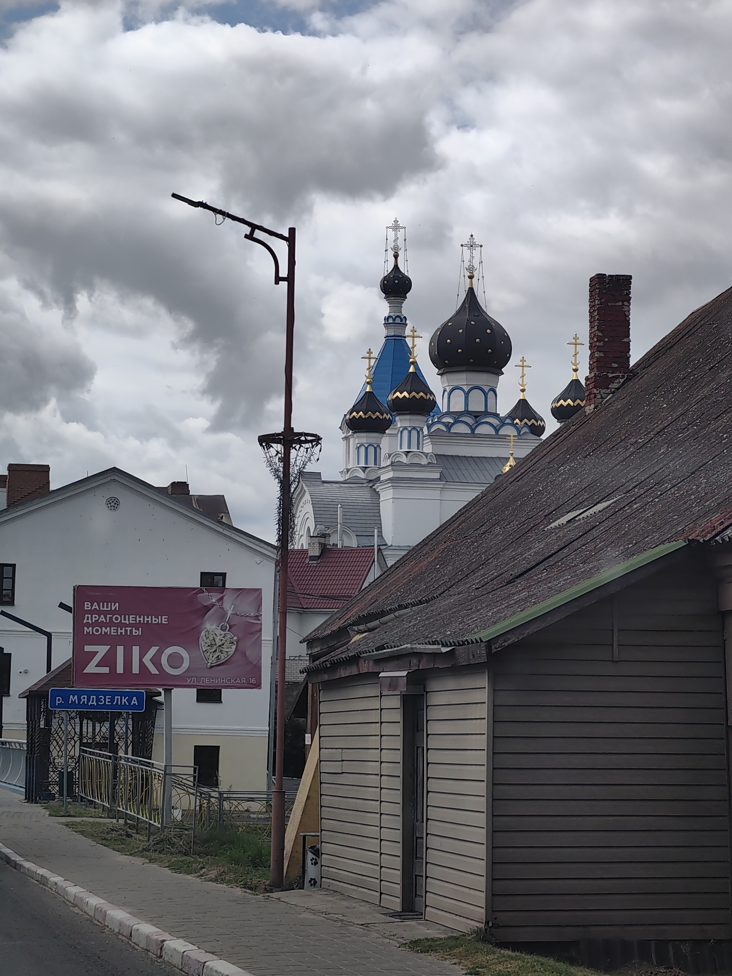 The image shows a street scene in Pastavy, Belarus, featuring a prominent church with multiple domes and crosses, a billboard advertising ZIKO jewelry, and a street sign indicating "r. MYADZELKA". The sky is overcast with heavy clouds. In the foreground, there is a sidewalk, a street, and a wooden building. The church's architecture is traditional Eastern Orthodox, with blue and black domes accented with gold. The billboard displays text in Cyrillic and an image of a heart-shaped pendant. The street sign is blue with white lettering. The overall atmosphere is somewhat somber due to the cloudy weather.