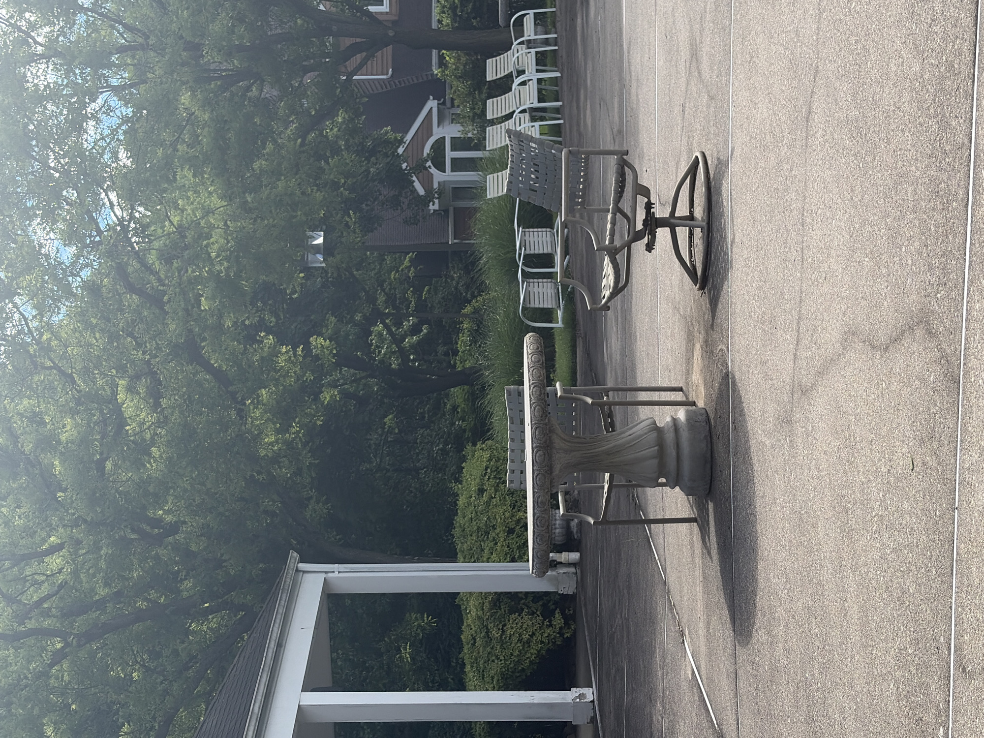 This image depicts an outdoor patio area in Edison, New Jersey, United States. The scene includes a concrete patio surface, a white stone table with decorative trim and metal legs, and a metal patio chair with a swivel base. In the background, there are several white lounge chairs, a glimpse of a building with dormer windows, and dense foliage from trees and shrubs. A white porch or patio structure with supporting columns is also visible. The lighting suggests it is daytime.
