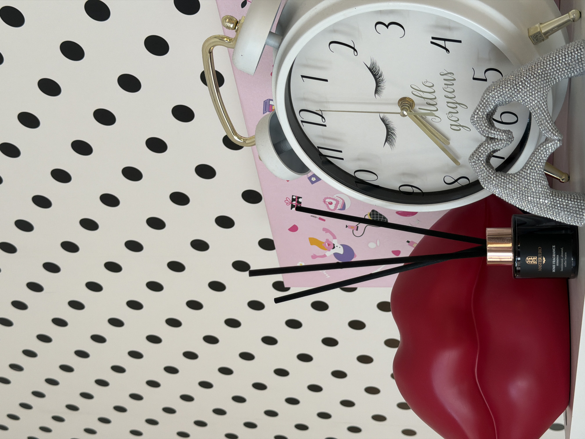 The image shows a still life composition, likely arranged for a photograph. The background is a white surface with a repeating pattern of black polka dots. Dominating the scene is a white alarm clock with black numerals and "Hello gorgeous" written on its face. The clock features gold-colored accents on the handle and bells. To the right of the clock is a silver-colored decorative object that resembles a heart. Below the clock, there is a pink box or notebook. A red, stylized lip-shaped container is positioned at the bottom of the image, with a black fragrance diffuser bottle nestled beside it. The diffuser includes several black reeds protruding from the bottle. The fragrance bottle is labeled "MASTERJERO HOME FRAGRANCE" with the scent "HERMINIA" specified as sandalwood, almond, and coffee beans. There is no indication of the location of Riga, Latvia, in the image.