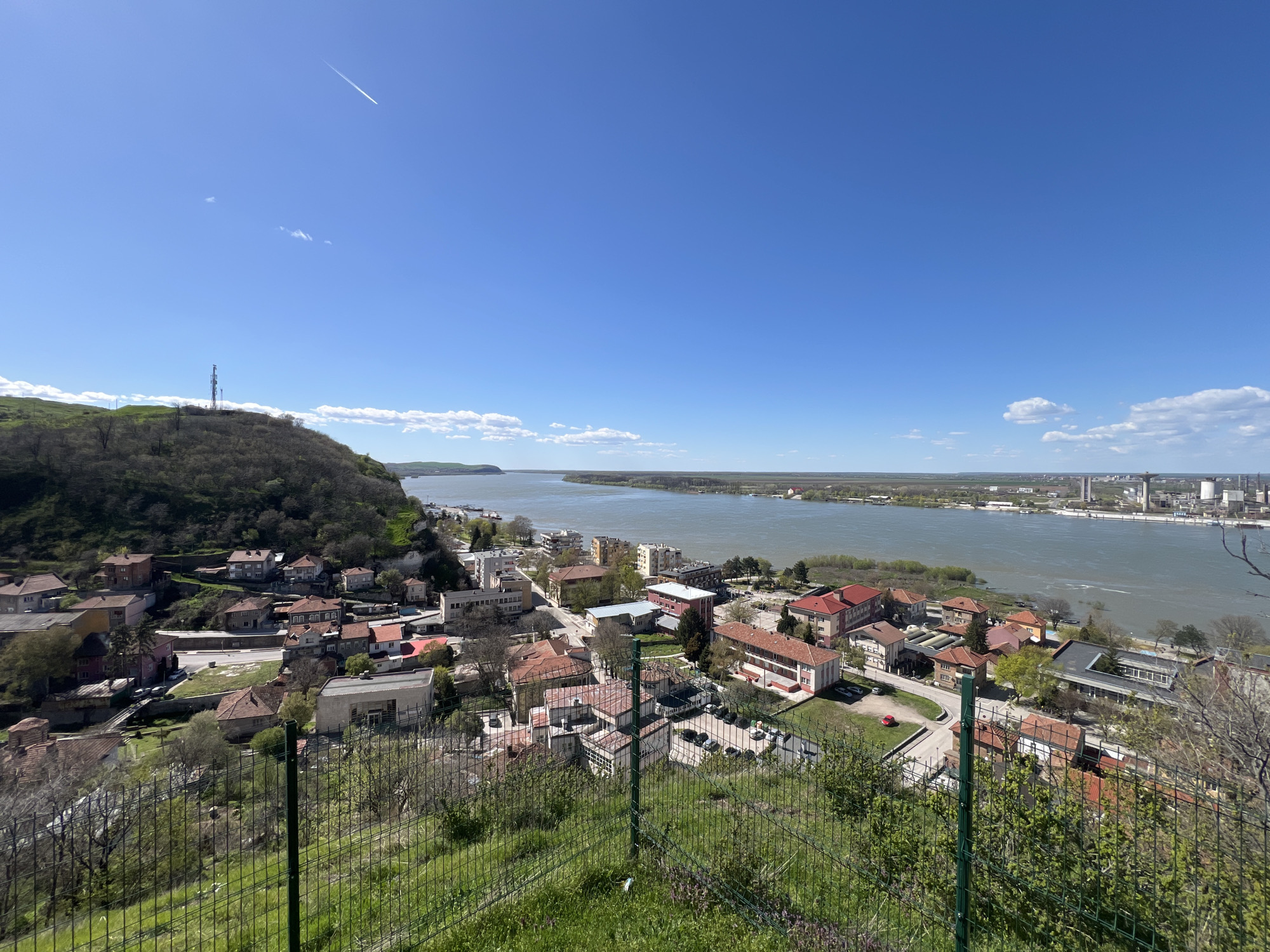 The image presents a high-angle view of a section of Nikopol, Bulgaria. The landscape comprises a mix of residential buildings with red-tiled roofs and modern structures, clustered near the shoreline of what appears to be the Danube River. The river spans the midground, stretching towards the horizon under a clear blue sky with minimal cloud cover. A hill with a communication tower rises on the left side of the frame, covered in trees. In the foreground, a metal fence separates the viewpoint from a grassy slope, suggesting the vantage point is an elevated area overlooking the town. The right side of the image shows industrial facilities along the riverbank. A condensation trail of a high-flying aircraft is visible in the sky.