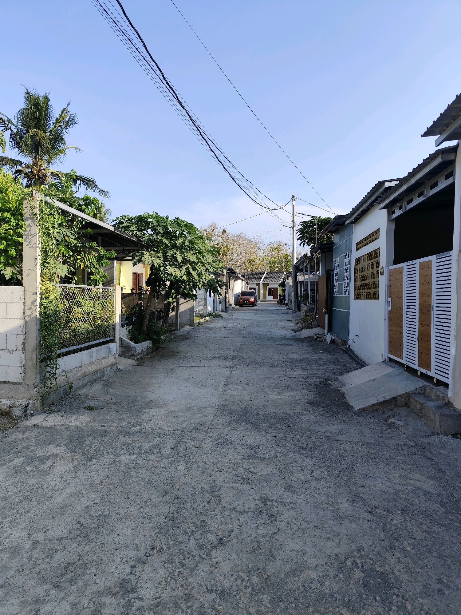 This is a vertical, eye-level, medium shot of a residential street in the city of Bambanglipuro, Indonesia. The street is paved with concrete and is lined with single-story houses on both sides. The houses appear to be modern and well-maintained, with pitched roofs and concrete walls. Some of the houses have open garages or carports. There are trees and vegetation along the street, and power lines are visible overhead. The sky is clear and blue, and the sun is shining. The scene appears to be quiet and peaceful, with no people or activity visible. The overall impression is of a typical residential neighborhood in a tropical climate.