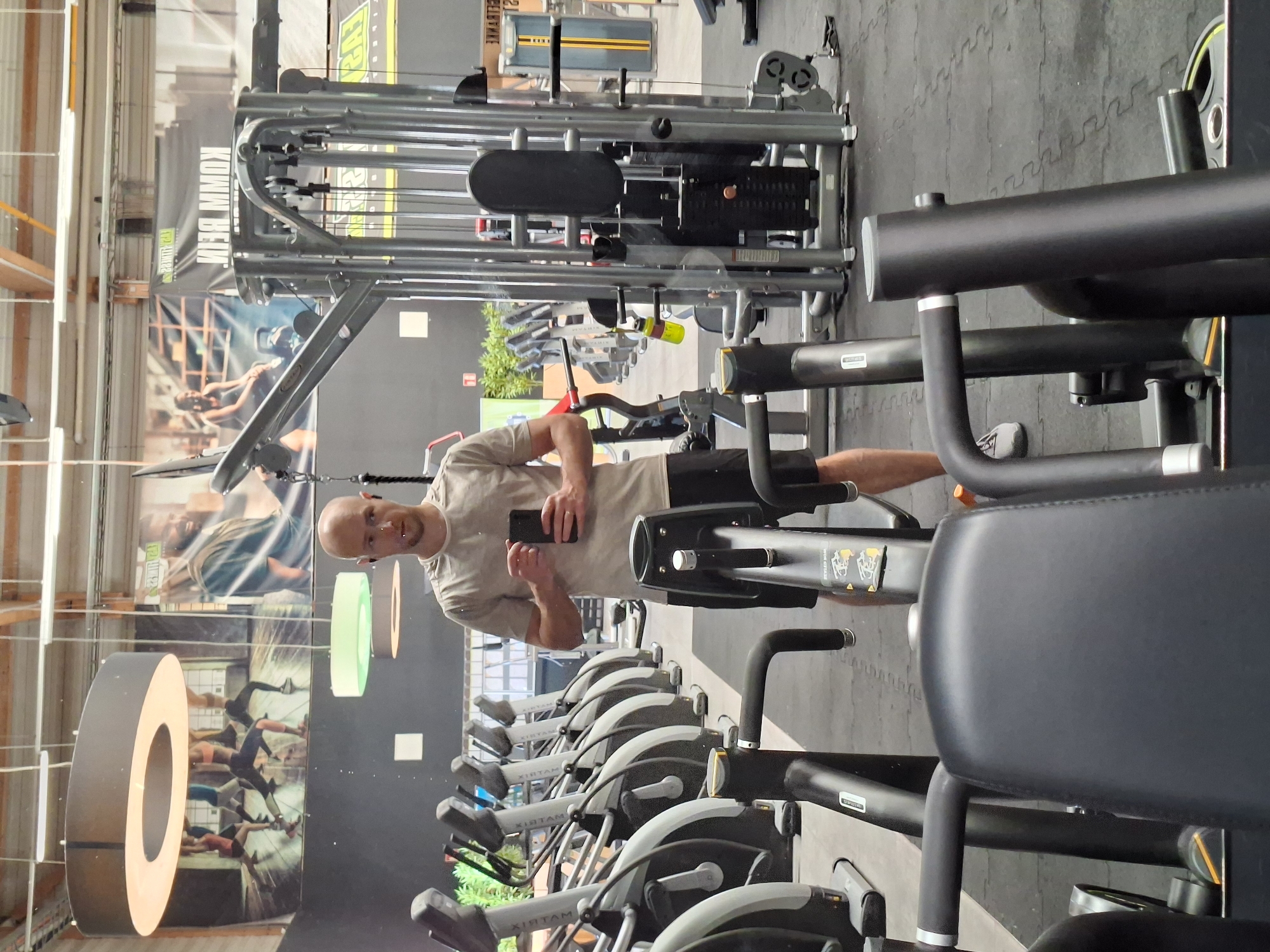 The image captures a full-length mirror reflection of a bald man in a gym located in Baden-Baden, Germany. The man is wearing a light grey t-shirt and black shorts, holding a smartphone in his right hand. He is standing in front of a row of elliptical machines and a weight machine. The gym environment is evident with the presence of various exercise equipment, large posters on the walls, and industrial-style lighting fixtures. The floor is covered with black rubber mats. The overall scene suggests a typical gym setting with a focus on fitness equipment and a workout atmosphere.