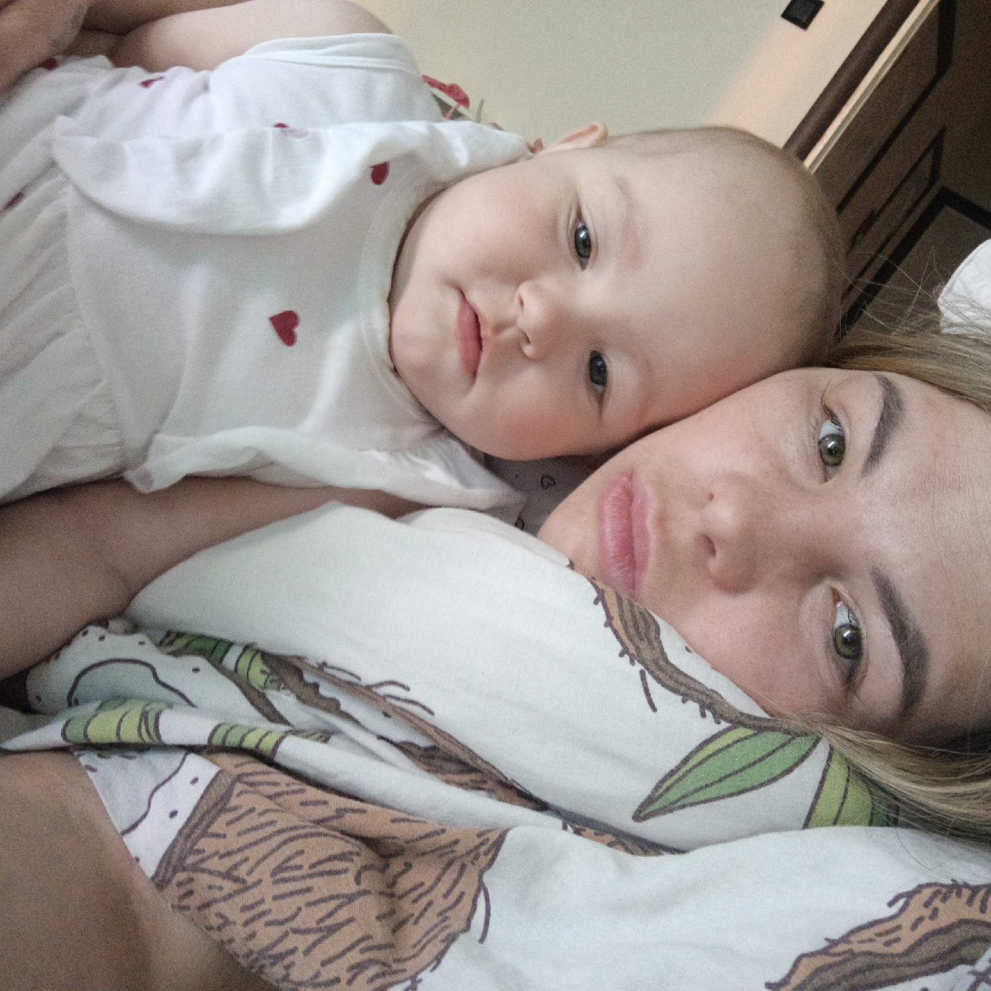 A woman and baby are lying on a bed, appearing to be indoors. The baby, wearing a white dress with small red hearts, is positioned above the woman, resting its head near hers. The woman is looking directly at the camera with her head on a pillow or blanket that has a pattern of what appears to be leaves and stylized animals in brown and green on a white background. Her blonde hair is visible near her face. A doorway or dark piece of furniture is partially visible in the upper right background.