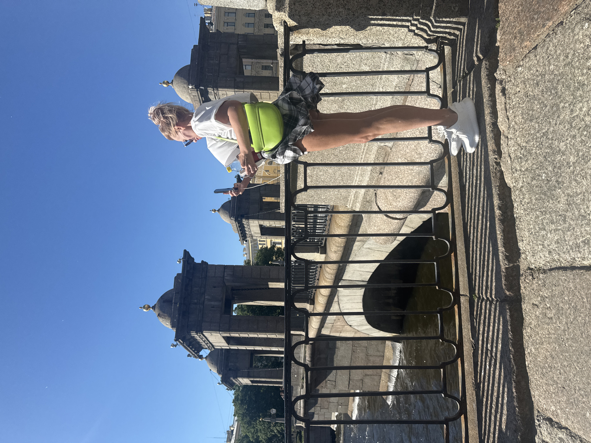 A blonde woman with her hair in a bun and wearing sunglasses, a white t-shirt, a plaid skirt, and white sneakers stands on a stone bridge in Saint Petersburg, Russia. She holds a smartphone in her right hand and a water bottle in her left. A bright green purse is slung over her left shoulder. The woman is positioned on the right side of the frame, facing left, with her profile visible.
Behind her, a stone bridge with decorative black metal railings spans a body of water. The bridge features arched stone supports and two symmetrical stone structures with domed roofs topped with golden finials. These structures appear to be part of the bridge's design, possibly housing mechanisms or decorative elements. The sky above is a clear, bright blue. Trees and buildings are visible in the background, suggesting an urban setting. The sunlight casts long shadows from the bridge railings onto the stone walkway where the woman is standing.