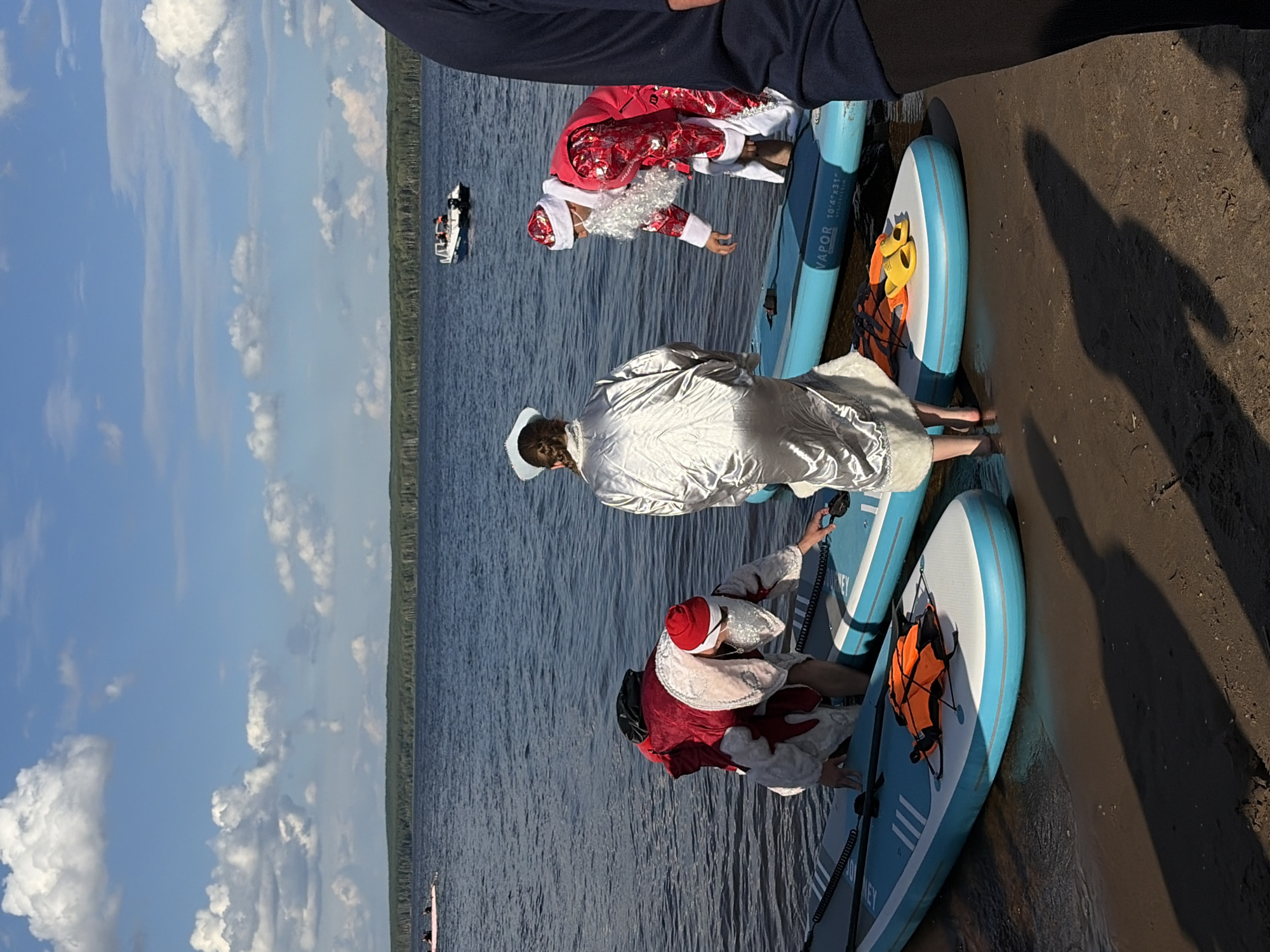 The image captures a scene at the Kama River embankment in Chaikovsky, Russia. The foreground shows a sandy shore with dark shadows cast by people. Several inflatable paddleboards are partially submerged in the water along the shoreline. On the paddleboards, three individuals are present. Two are dressed in Santa Claus costumes, one in a red suit with white trim and a red hat, and another in a red suit with white trim and a white beard. The third individual is wearing a silver, shiny robe and a white hat. An orange life vest is visible on one of the paddleboards. In the middle ground, the Kama River stretches out, with a few boats visible in the distance. The background features a dense, dark green forest line against a bright blue sky with scattered white clouds. The lighting suggests it is daytime, likely late afternoon or early evening due to the long shadows.