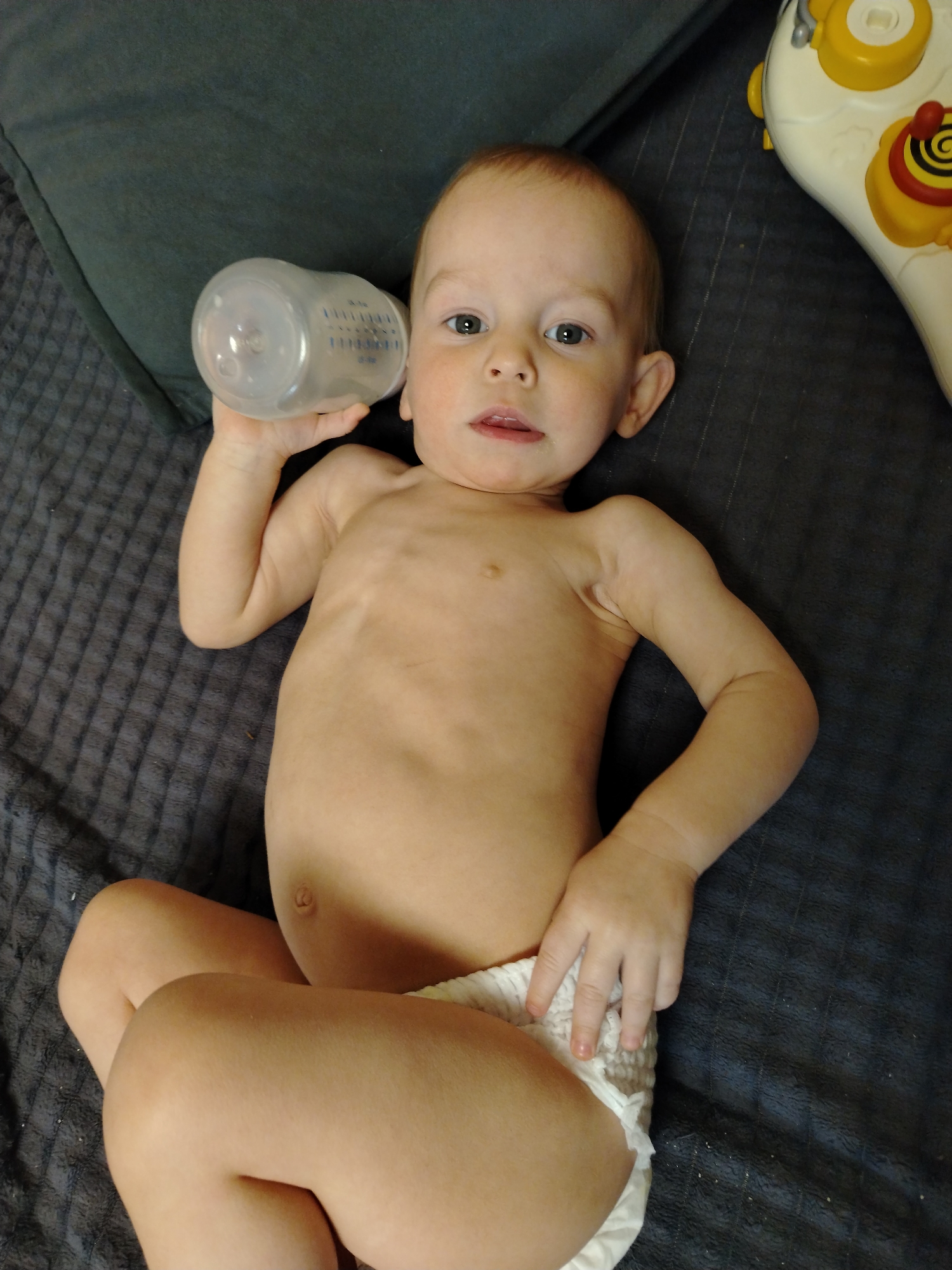 An infant is depicted lying supine on a dark grey, textured blanket surface in the city of Chaikovsky, Russia. The infant, wearing a white disposable diaper, has light brown hair and green/hazel eyes, and is looking directly towards the viewer with a slightly open mouth. Its right hand holds a clear plastic baby bottle with blue measurement markings, positioned near its head. A dark grey pillow is visible behind the infant's upper left side, and a white and yellow plastic toy with red accents is partially visible in the upper right background.